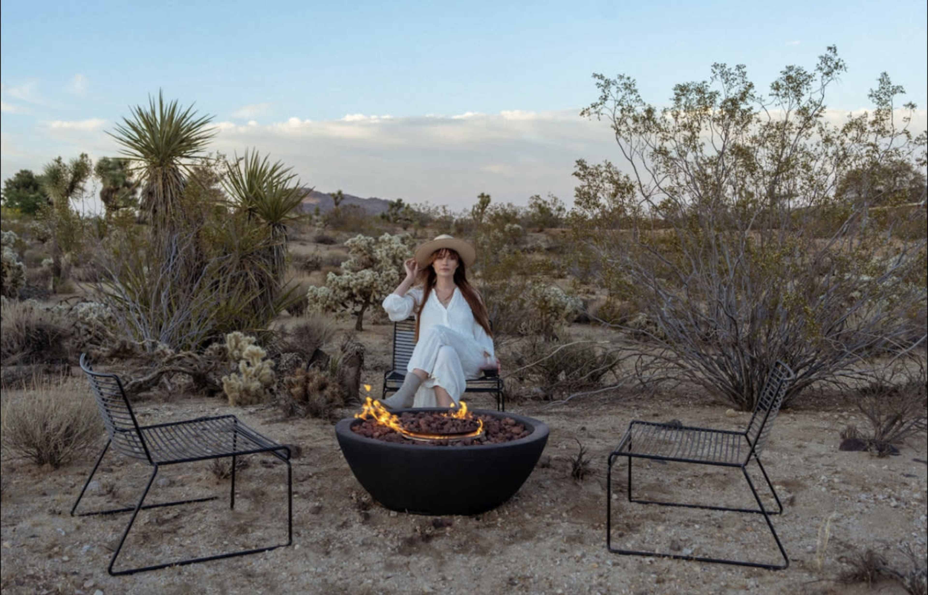 A woman in a wide-brimmed hat sits in a chair near a fire pit surrounded by desert vegetation and sparse bushes.