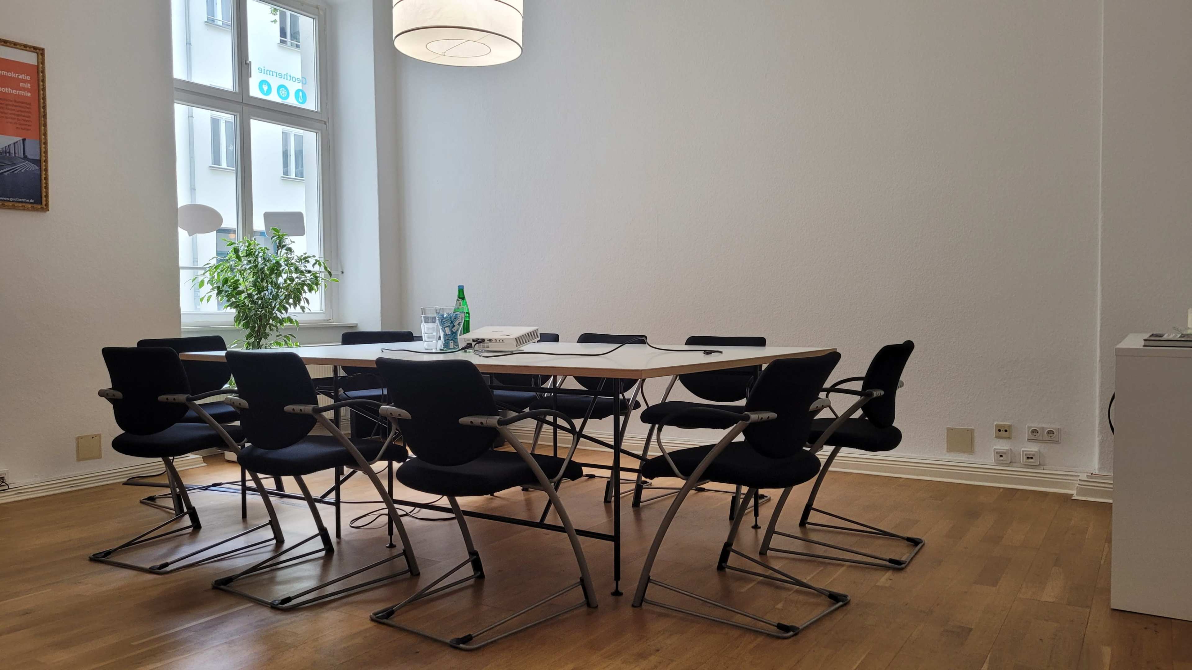 A large, minimalist conference room features a long table surrounded by eight black chairs, with a plant and water bottles on the table, and a bright window in the background.