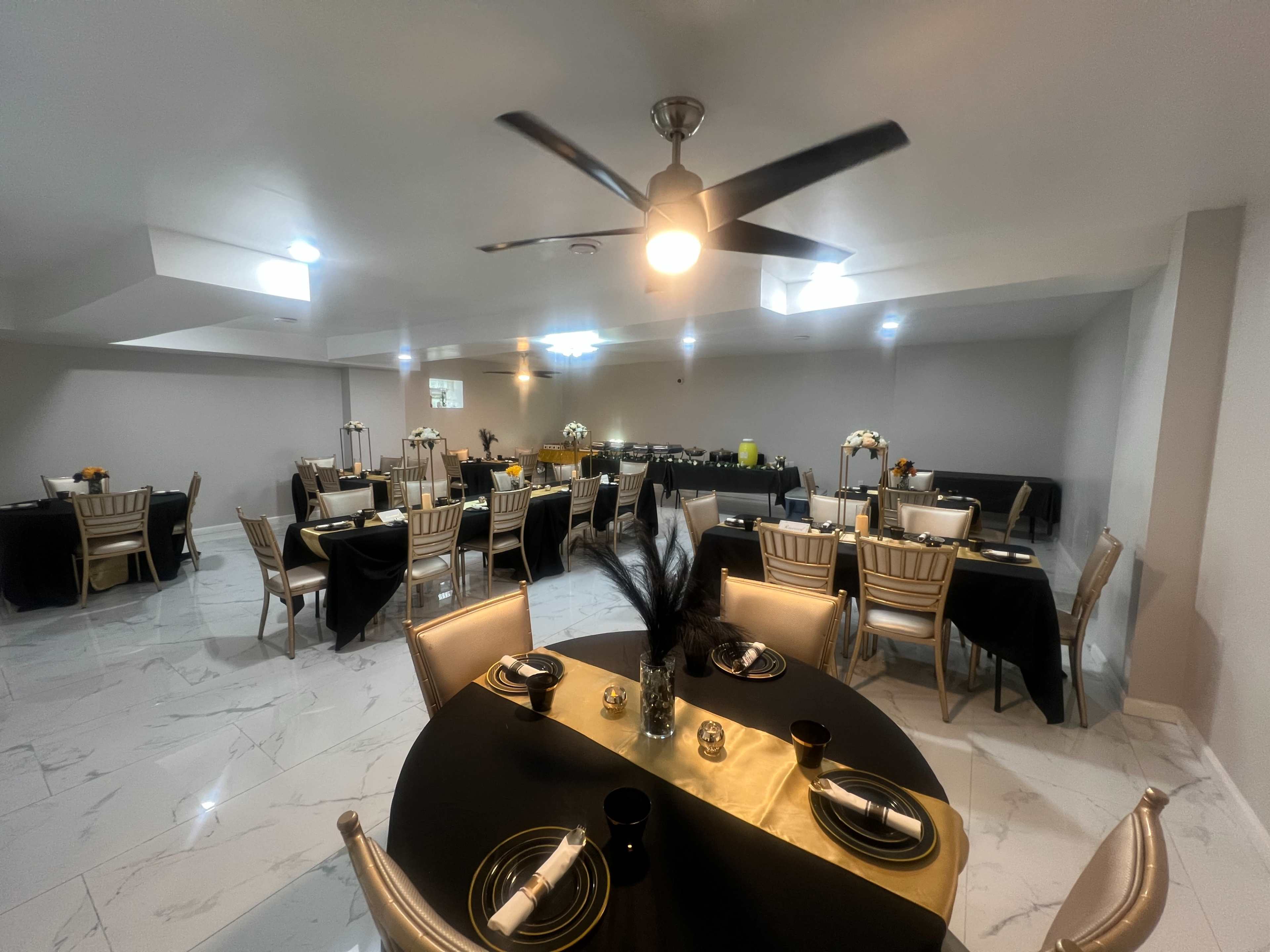 The image shows a spacious dining area set up for an event, featuring multiple tables with black and gold table settings, decorated with floral centerpieces.