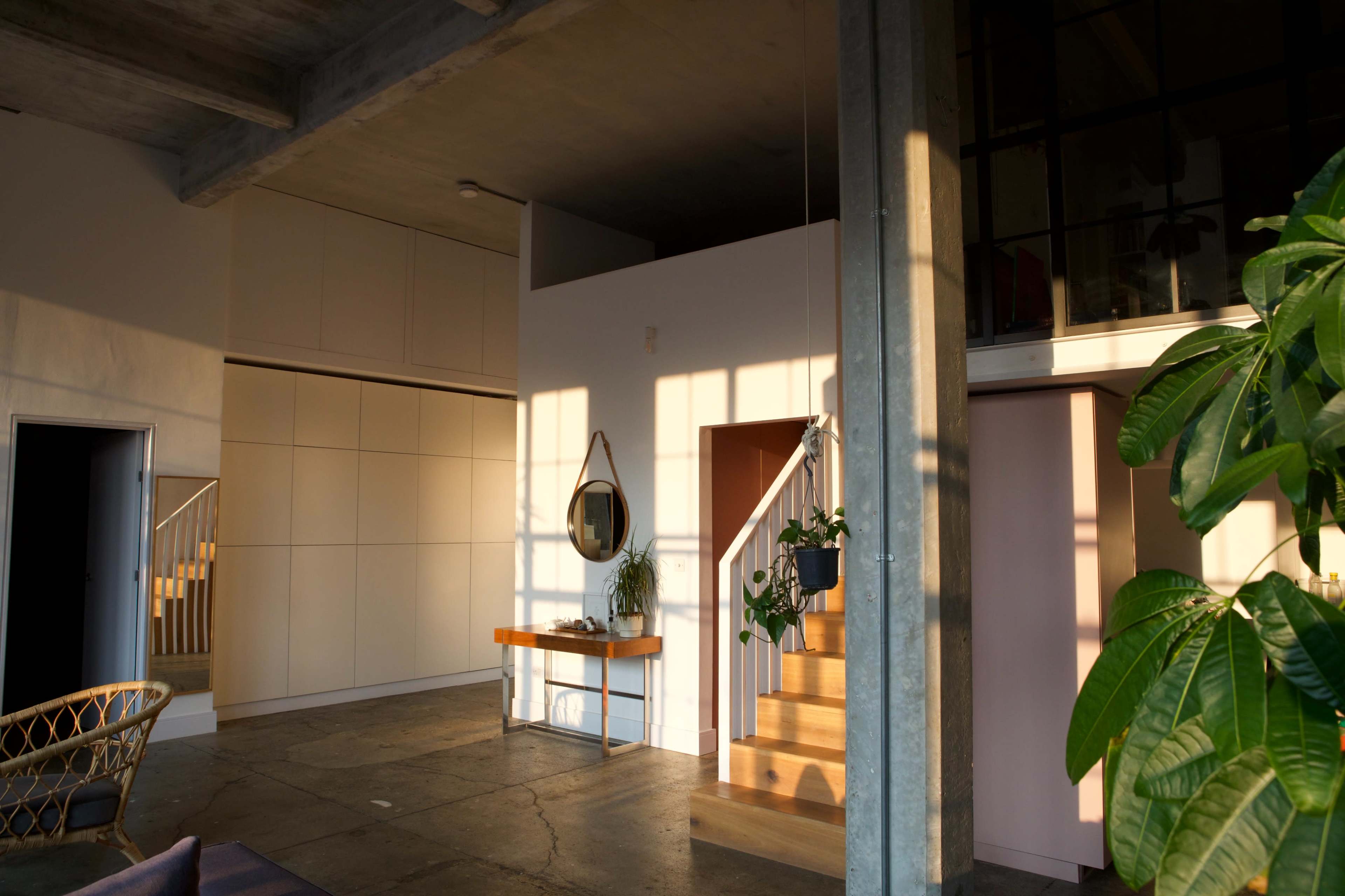 The image shows a modern interior space featuring a staircase, a mirror on the wall, and large windows allowing sunlight to fill the area.