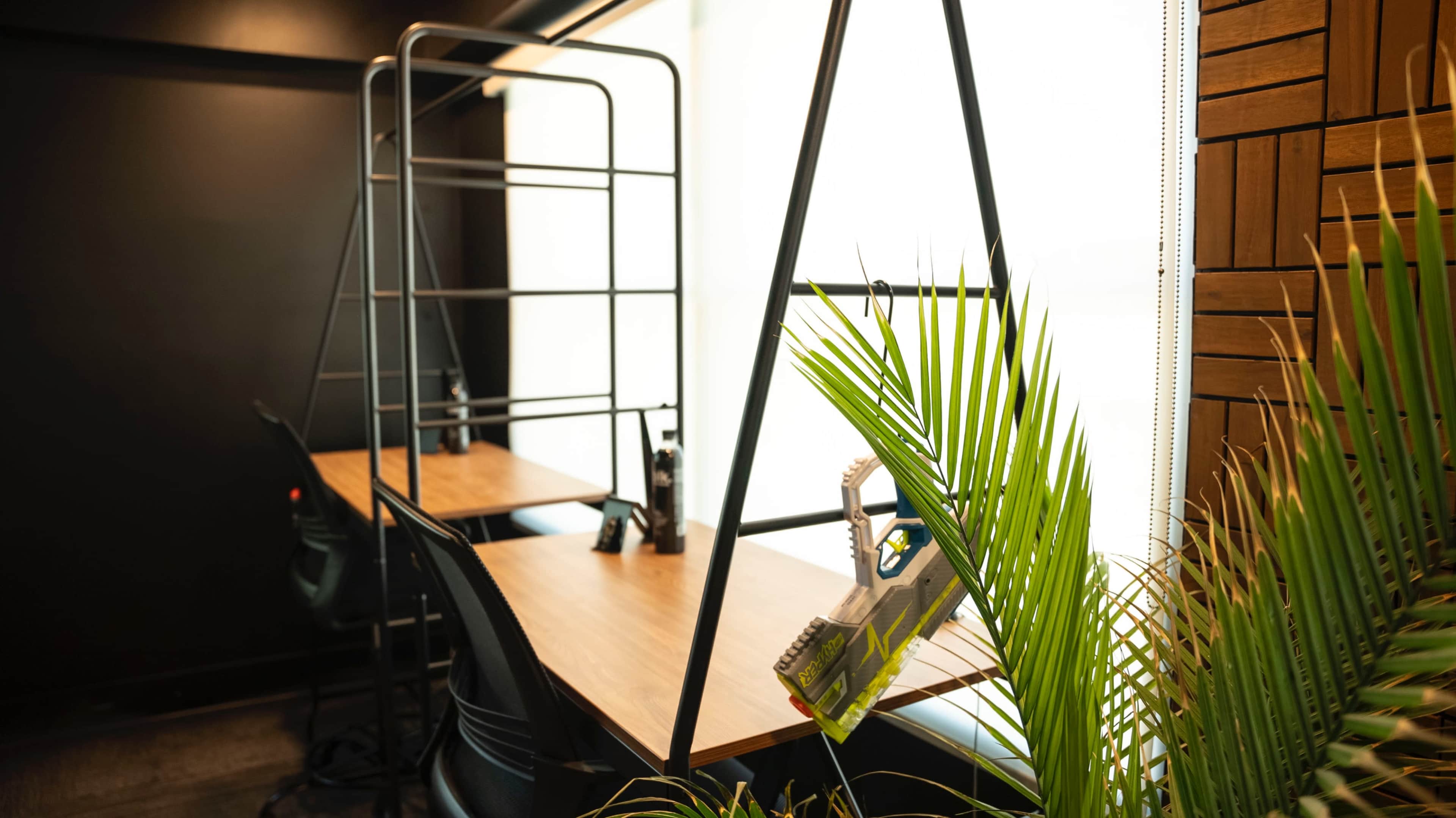 A minimalist workspace features two wooden tables with black chairs, surrounded by plants and a metal shelving unit near a large window.