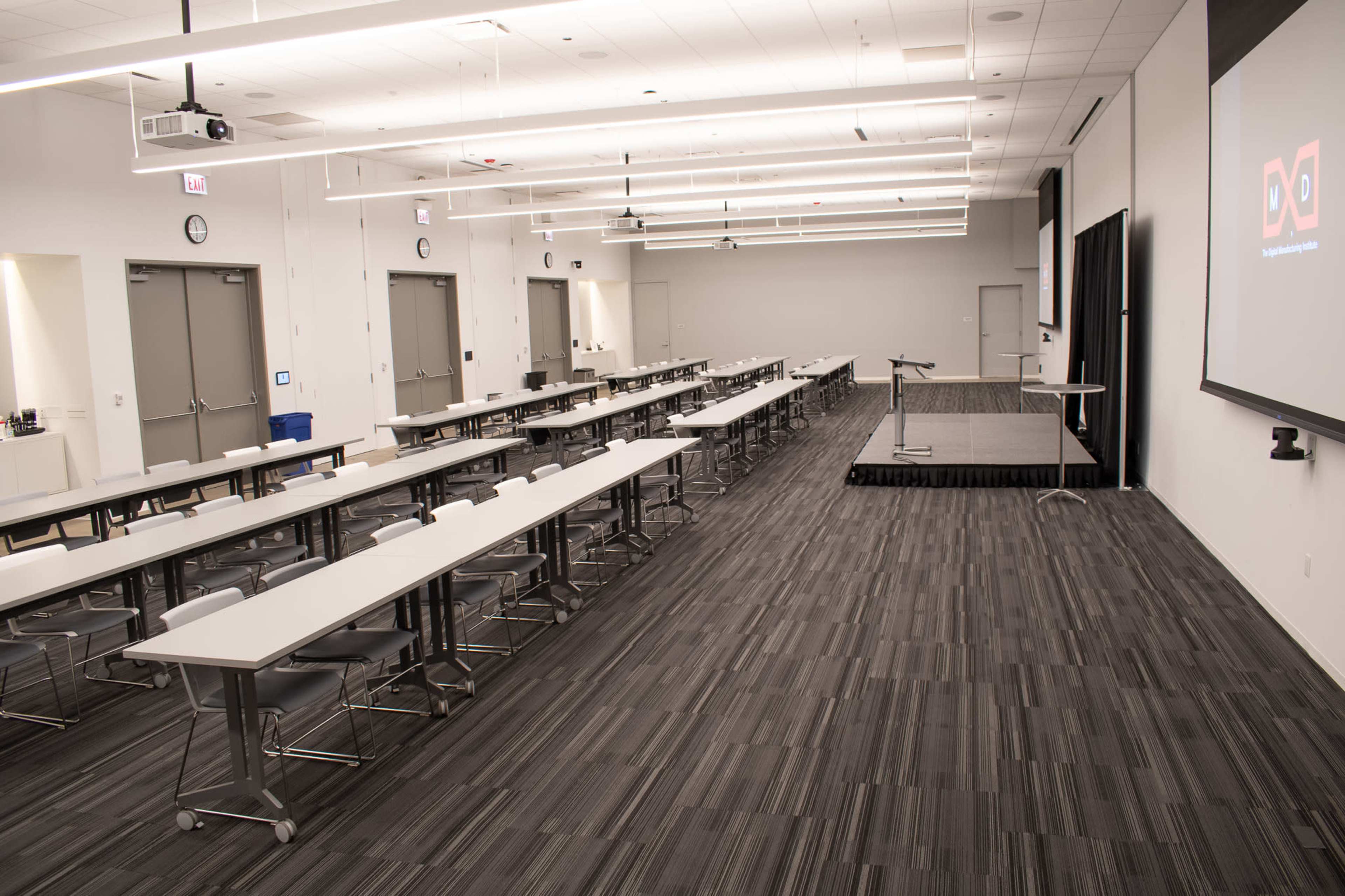 A spacious conference room features multiple long tables arranged in rows, with a podium and screen at the front, and clocks mounted on the walls.