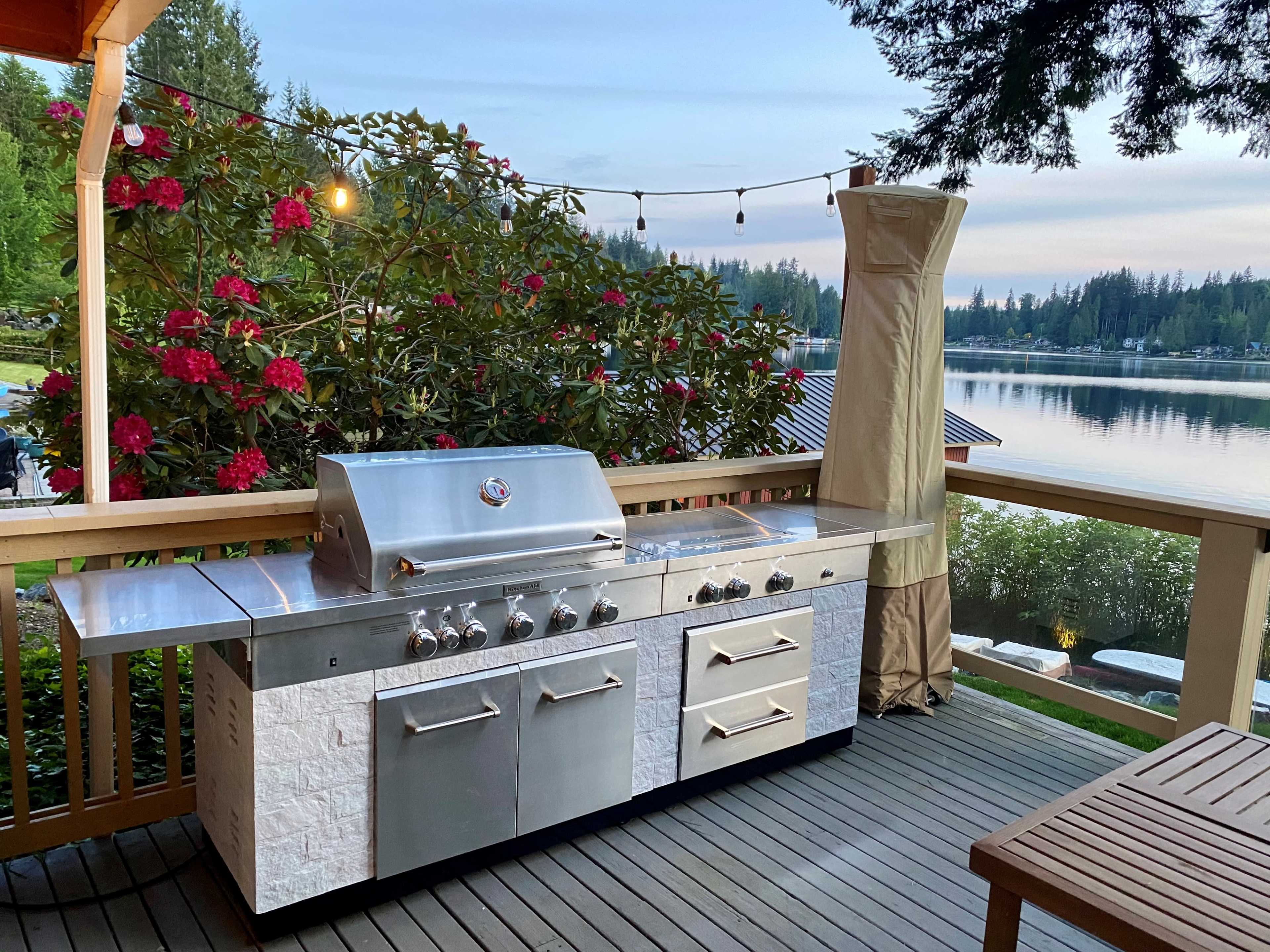 The image shows an outdoor kitchen featuring a stainless steel grill with a large countertop, situated on a deck overlooking a lake surrounded by trees and blooming flowers.