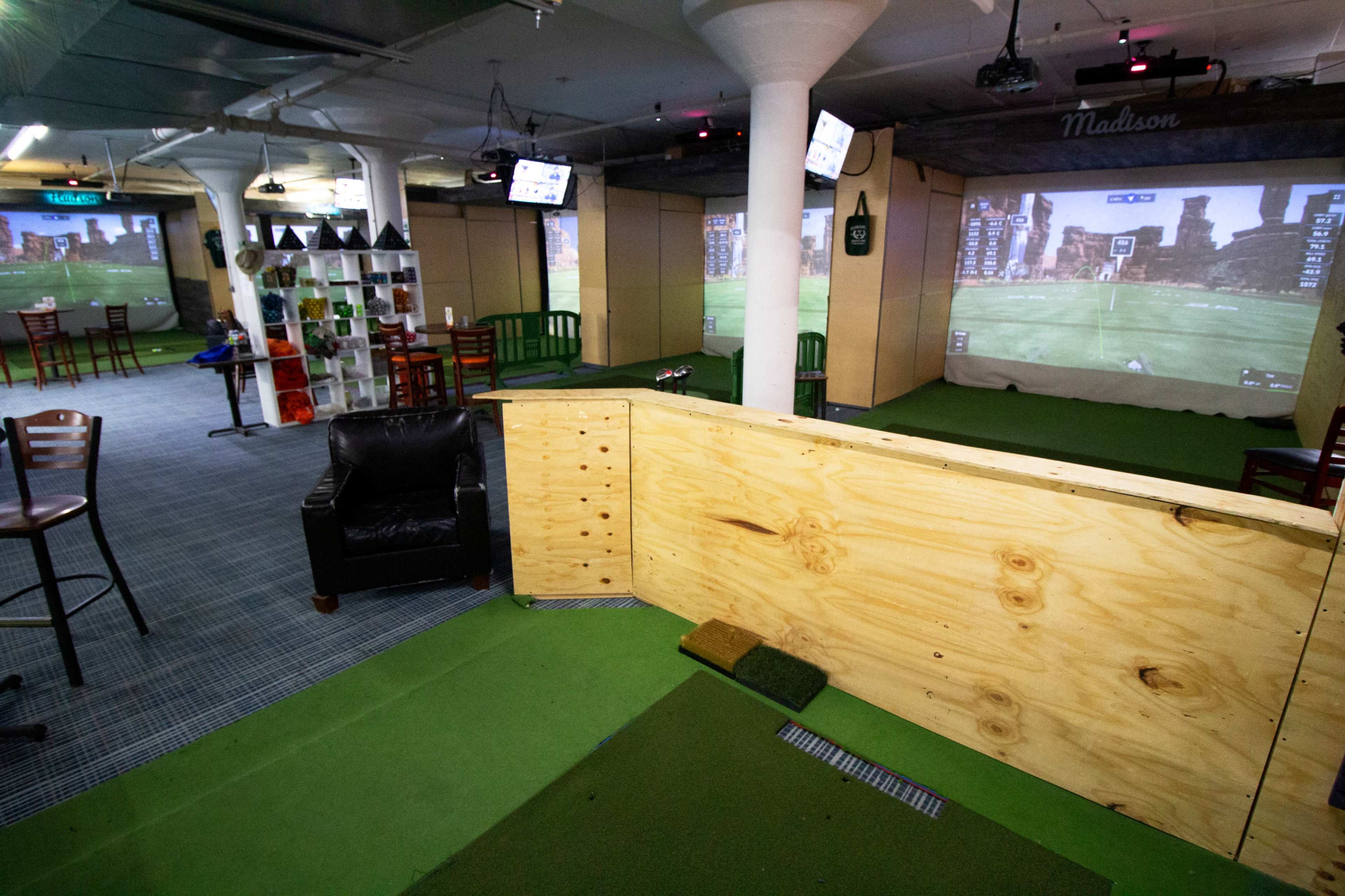 An indoor golf simulation area with a wooden barrier, artificial turf, and large screens displaying a golf course backdrop.