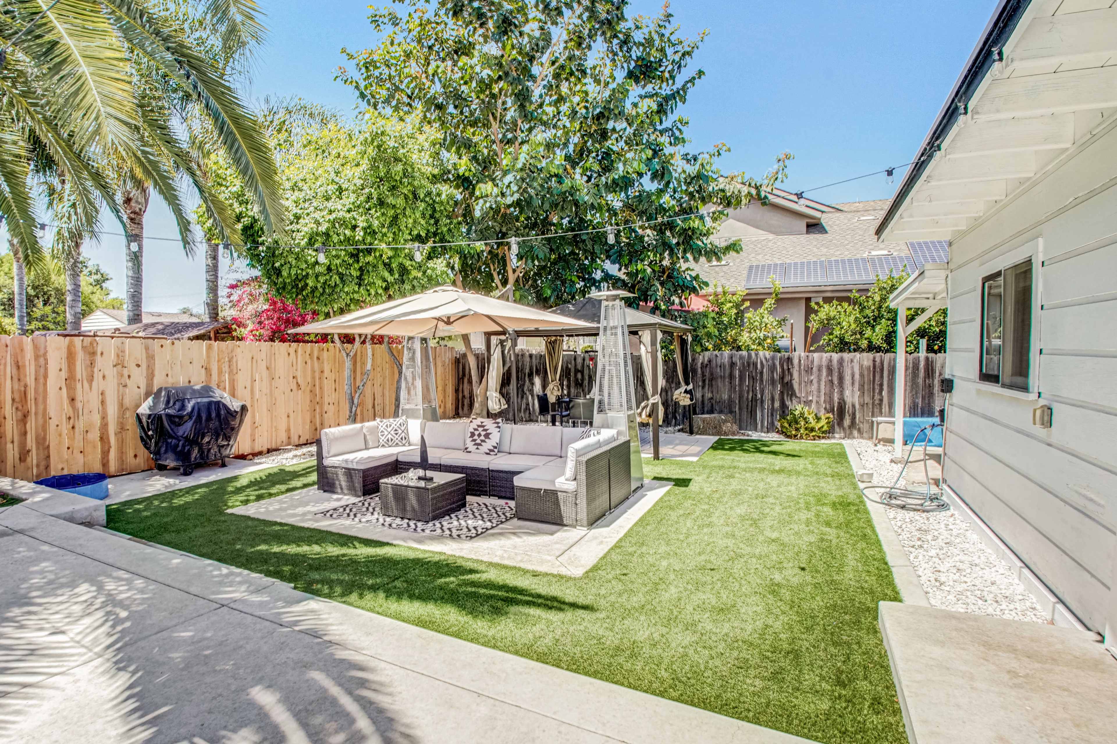 A backyard with a shaded seating area featuring a gazebo, surrounded by artificial grass and palm trees, with a barbecue grill in the corner.