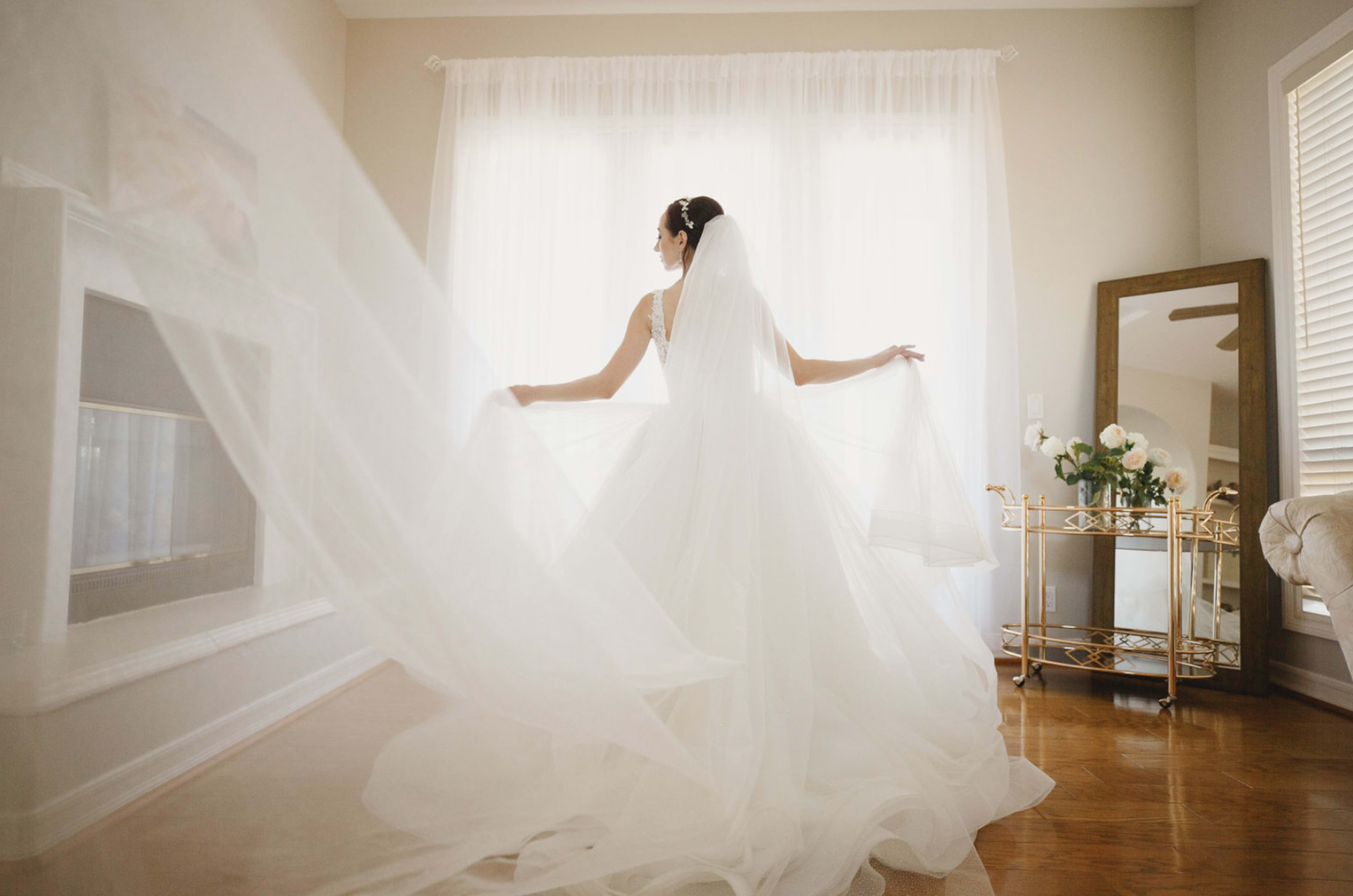 A woman in a wedding dress stands near a large window, holding her flowing veil and looking away from the camera.