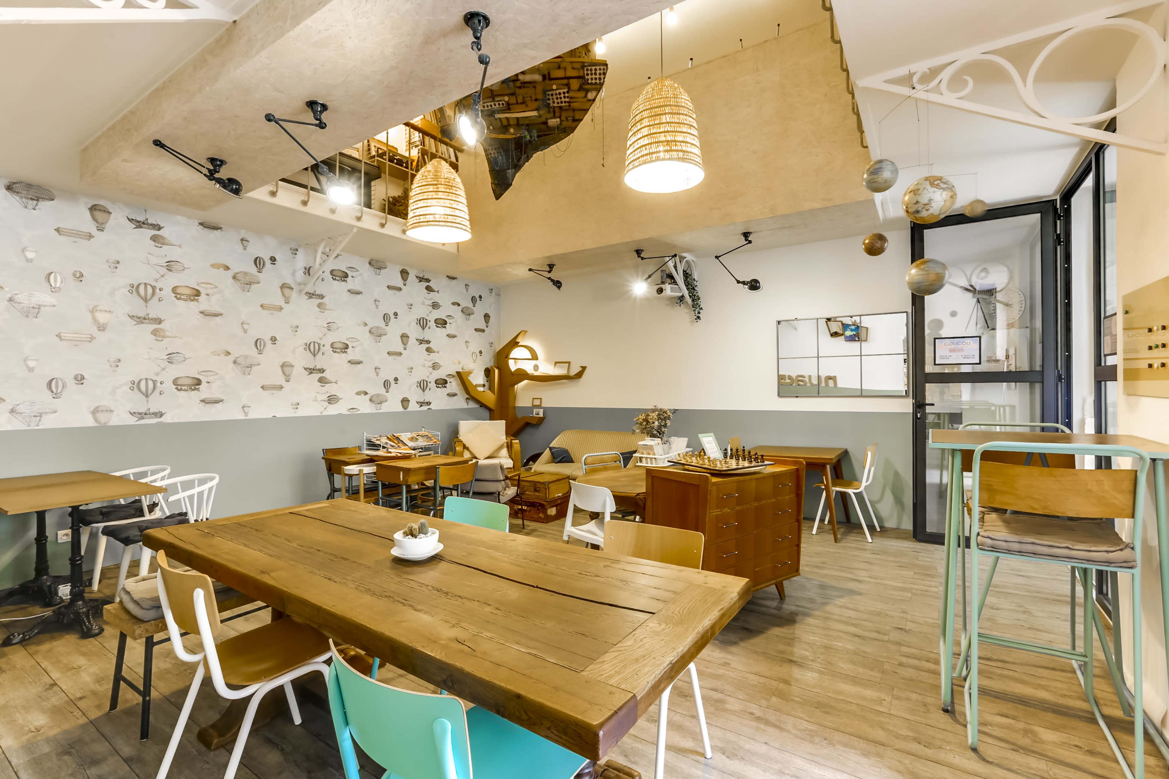 A cozy café interior featuring wooden tables, colorful chairs, and a staircase leading to an upper level decorated with books.