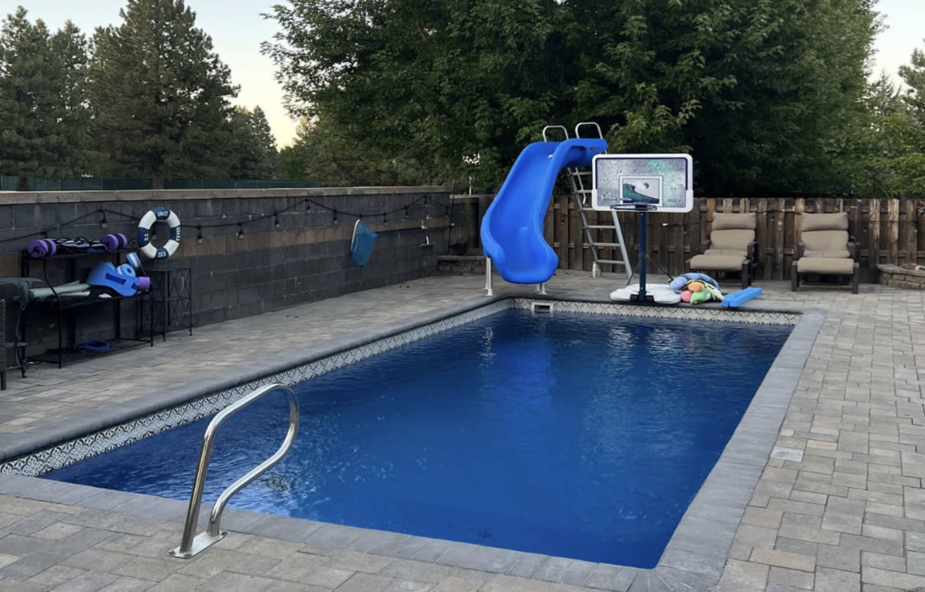 A backyard pool with a blue waterslide, a basketball hoop, and lounge chairs arranged beside the water.