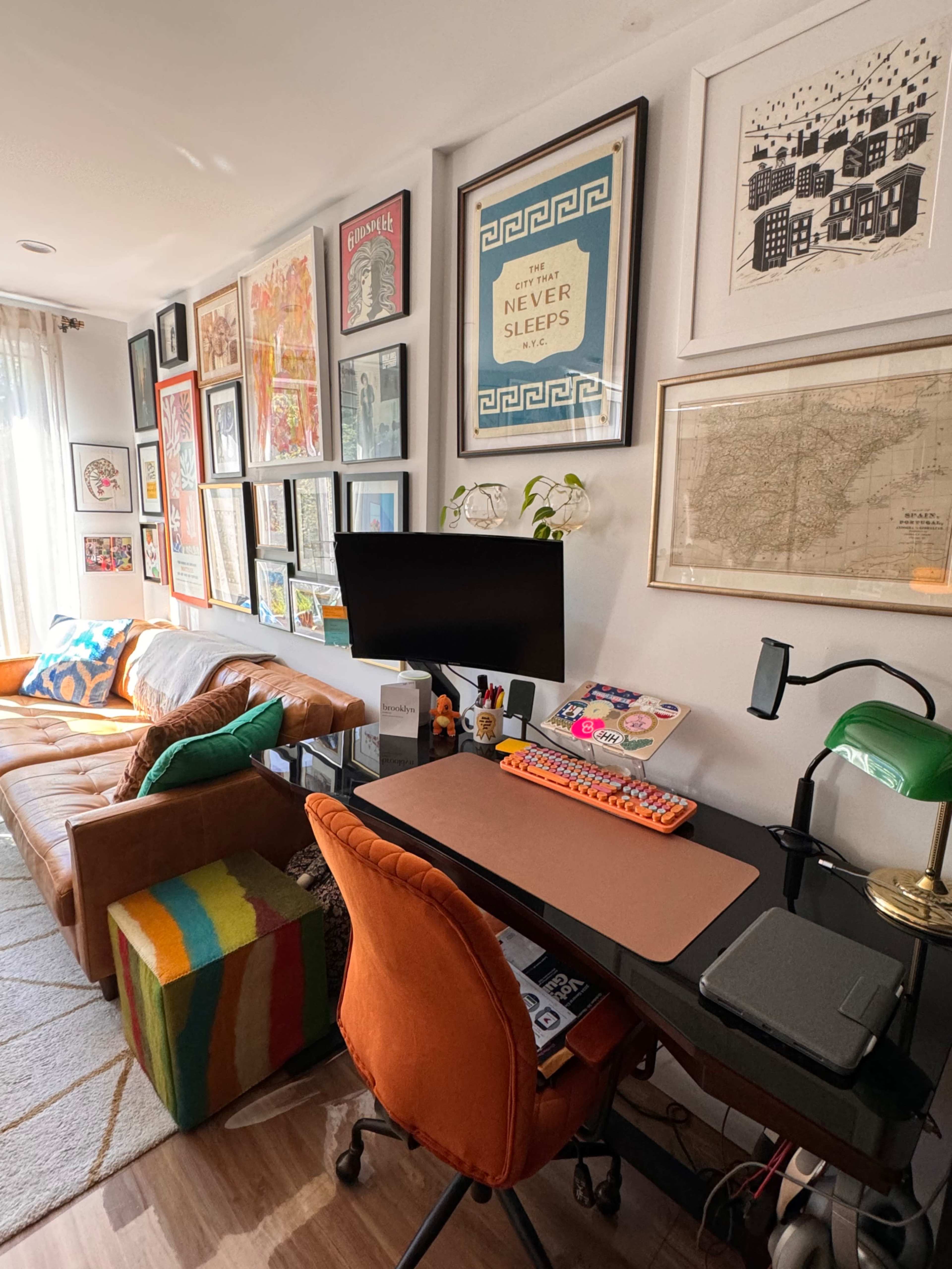 A colorful home office features a desk with a computer and various office supplies, surrounded by framed artwork and a couch with decorative pillows.