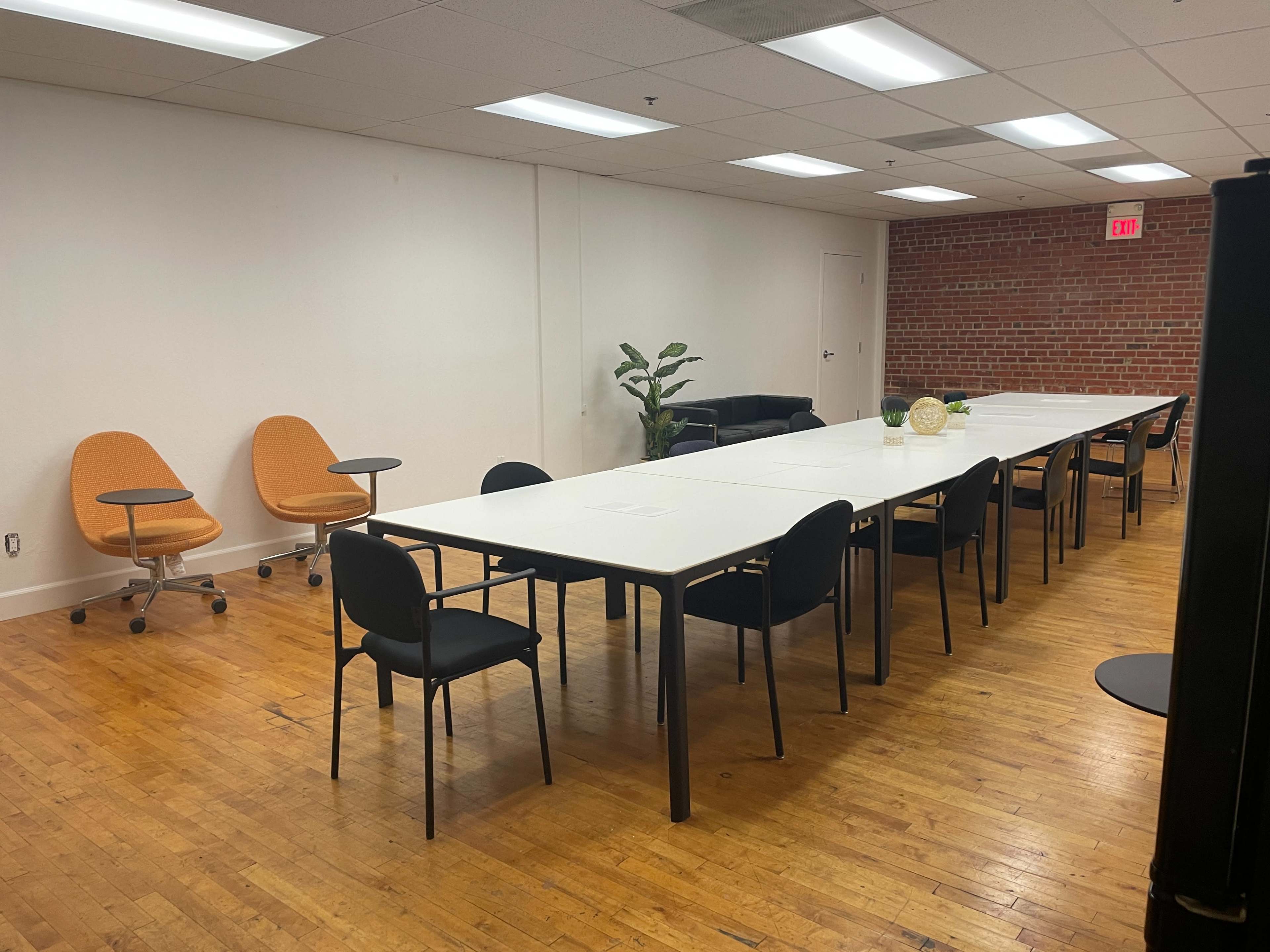 A long, rectangular table is surrounded by chairs in a well-lit meeting room with a brick wall and a sofa in one corner.