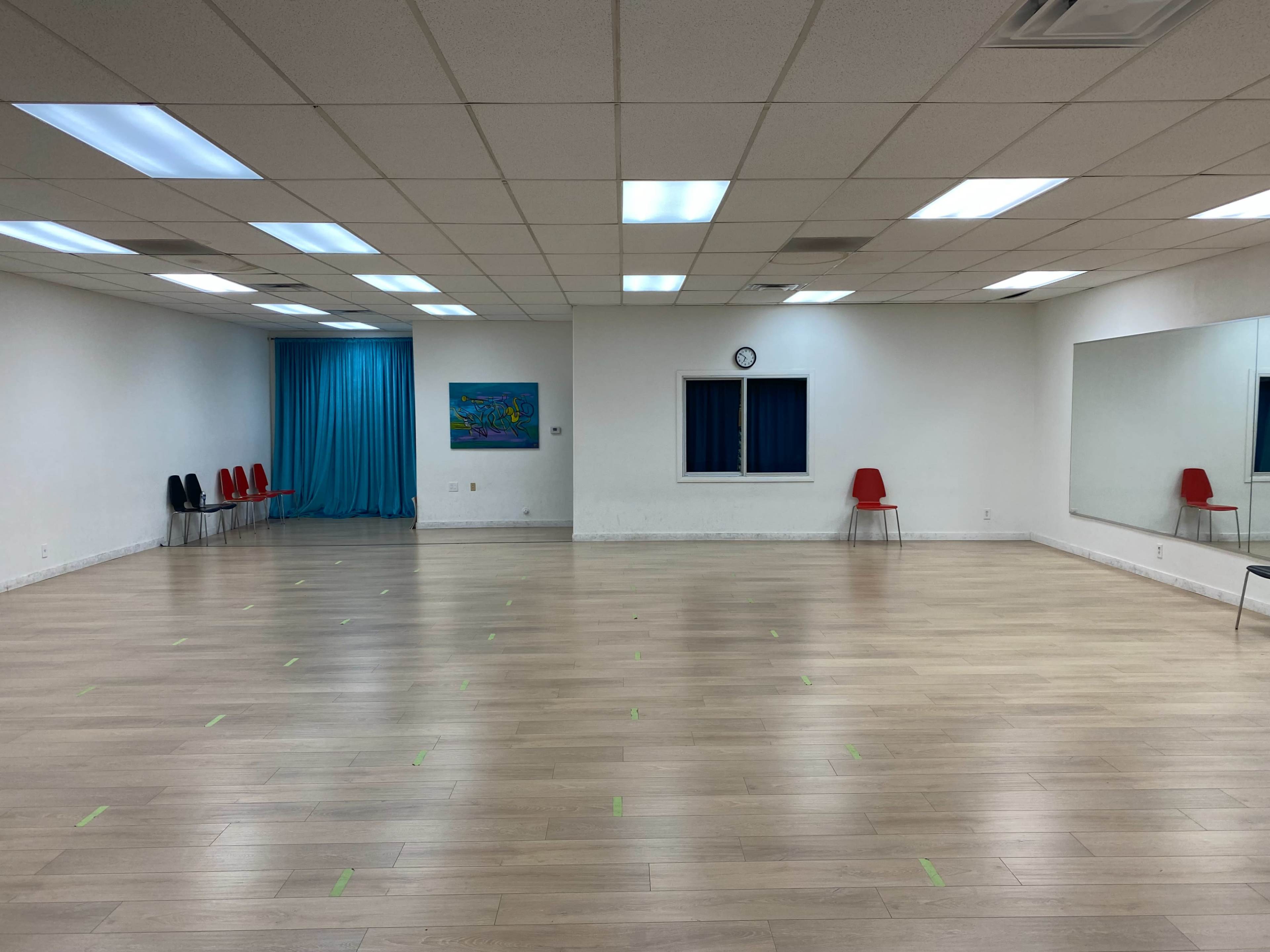 A spacious dance studio featuring a wooden floor, a large mirror, red chairs along the walls, and a blue curtain in the background.