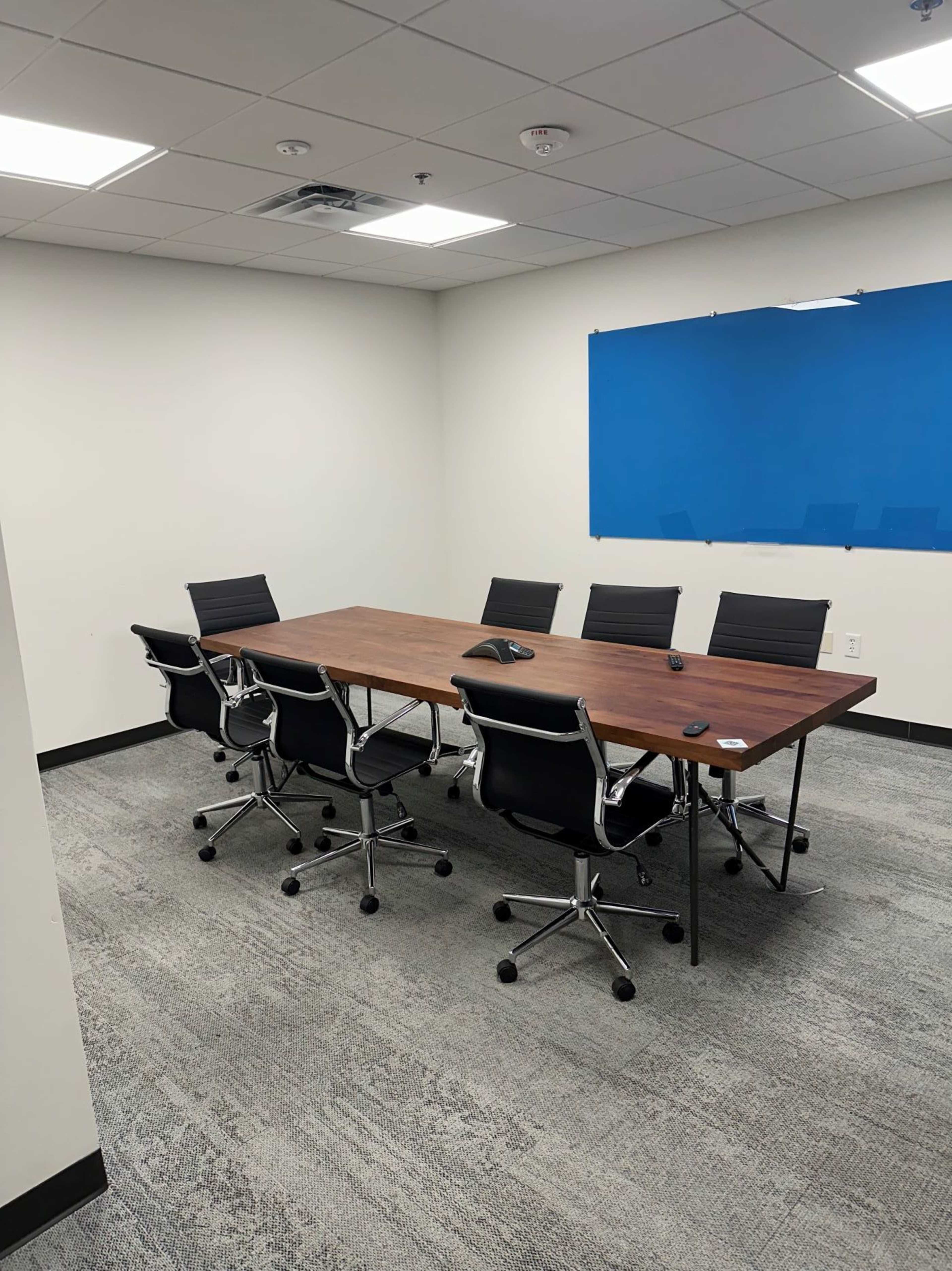 The image shows a conference room with a large wooden table surrounded by six black rolling chairs, and a blue wall in the background.