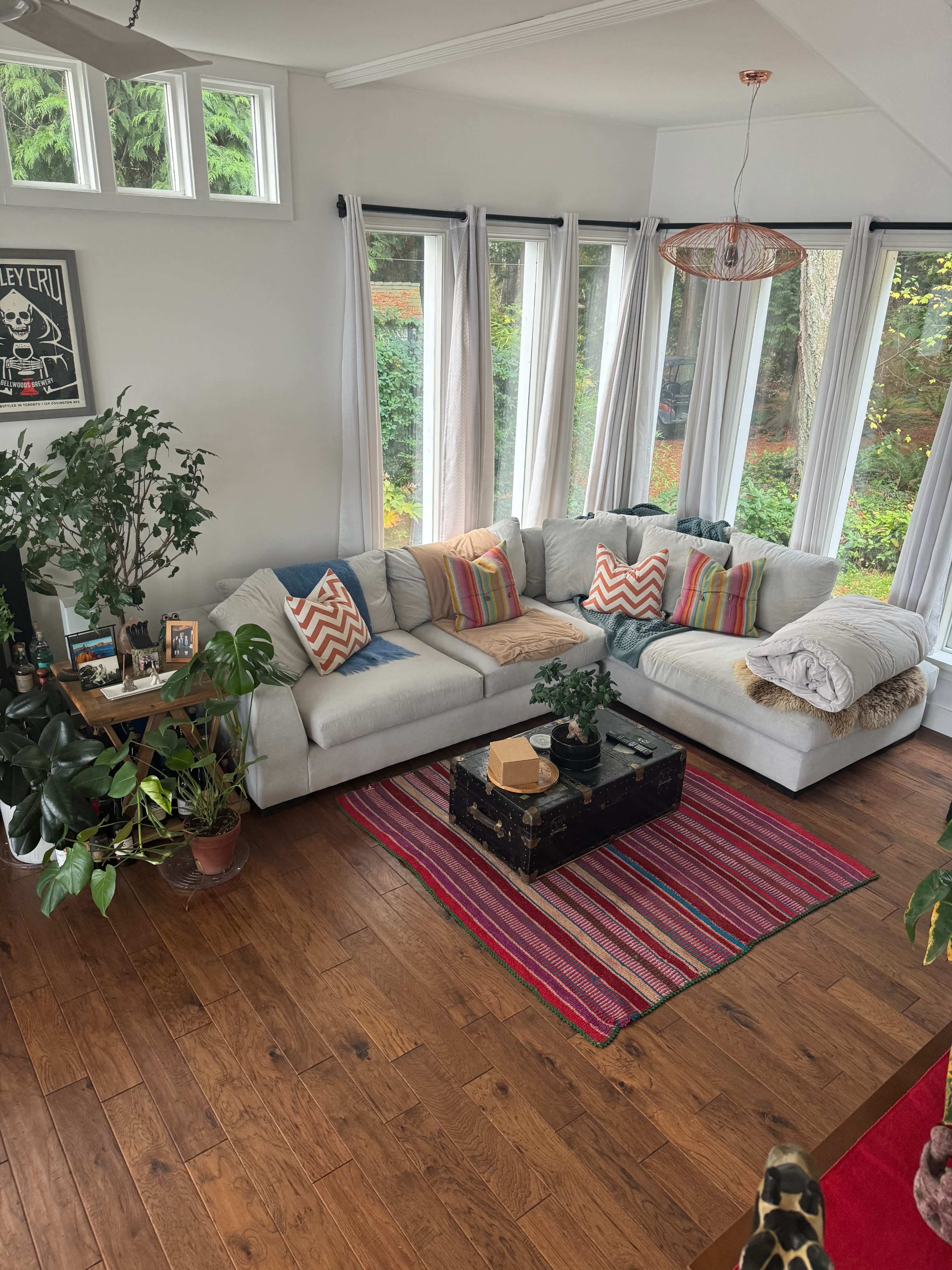 A bright living room with large windows, a white sectional sofa adorned with colorful pillows, a coffee table on a striped rug, and various plants and decor items.
