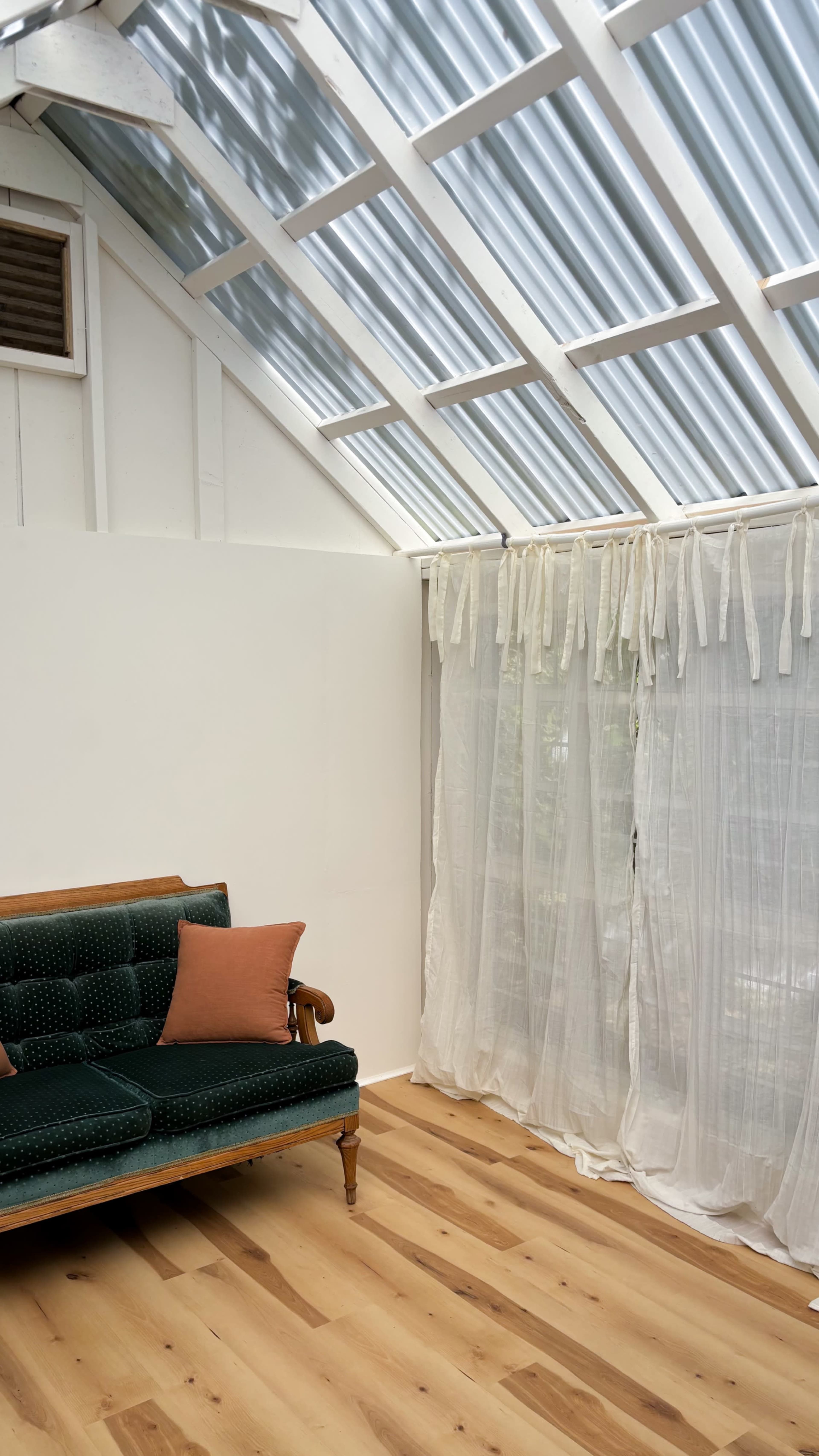 The image shows a bright room with a green sofa, two cushions, and sheer white curtains near a slanted glass roof.