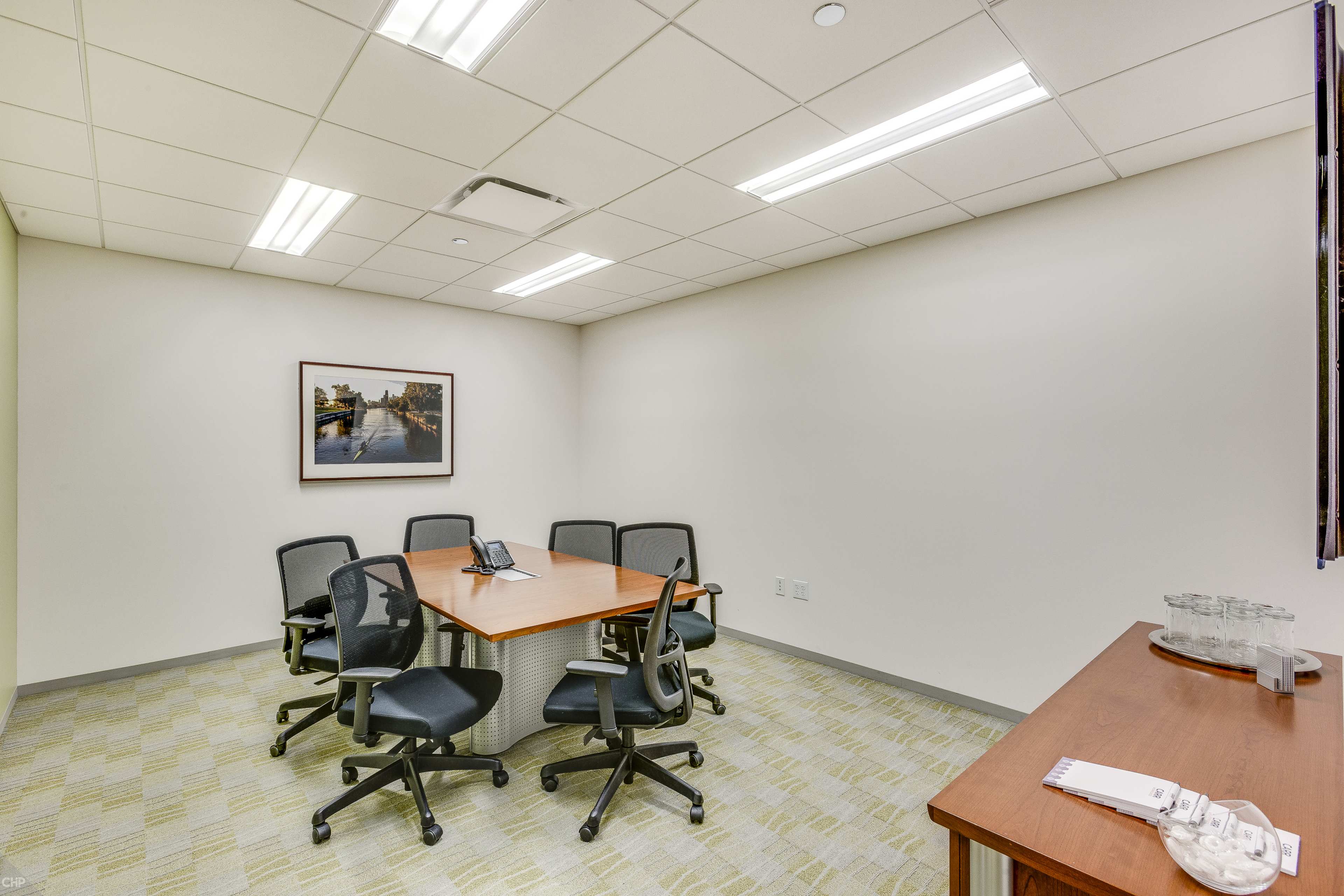 A conference room features a wooden table surrounded by six mesh office chairs, with a framed picture hanging on the wall and a side table holding glassware.