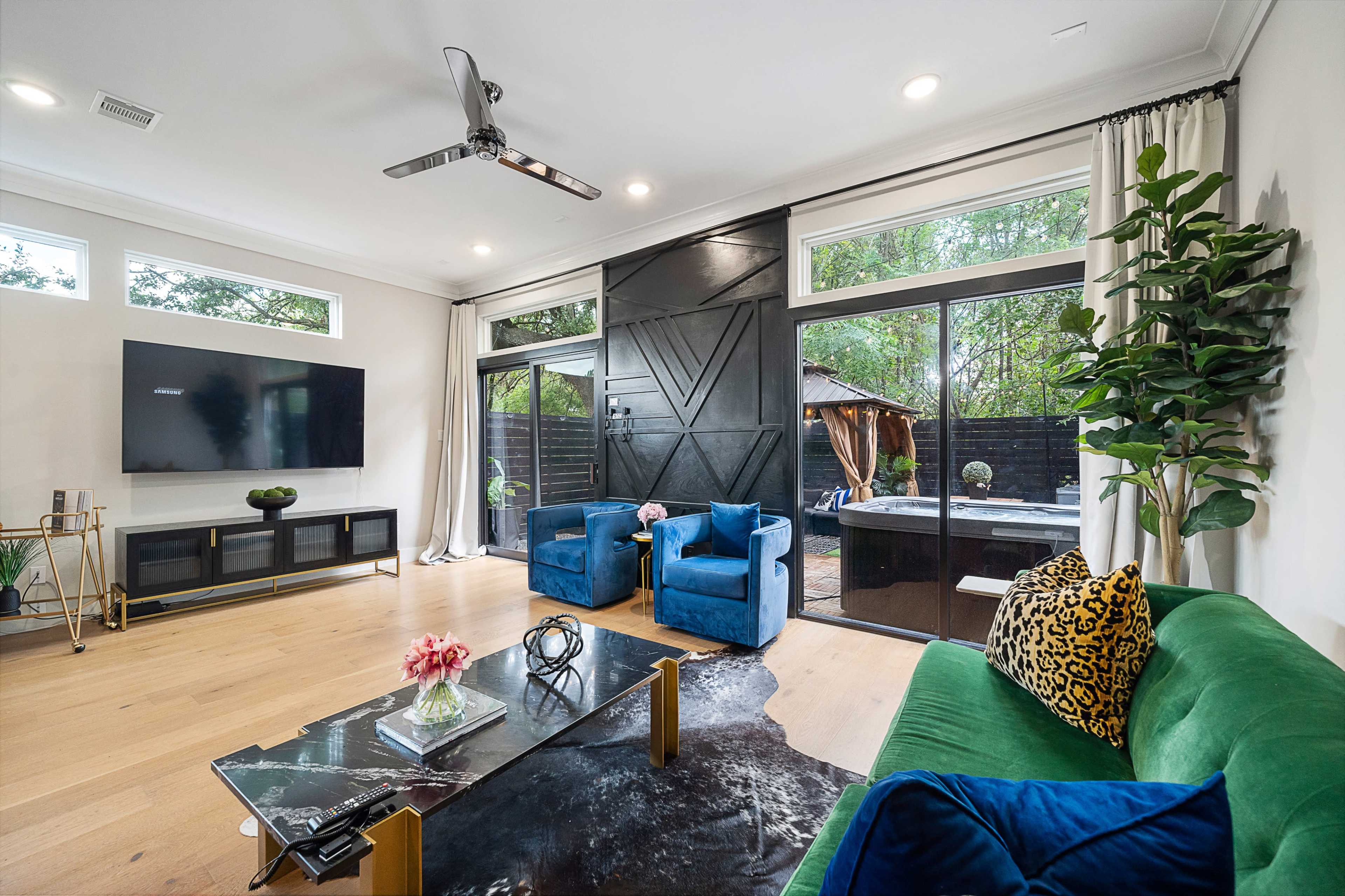 A modern living room with large windows, featuring a green sofa, blue chairs, a coffee table, and a wall-mounted television, surrounded by indoor plants.