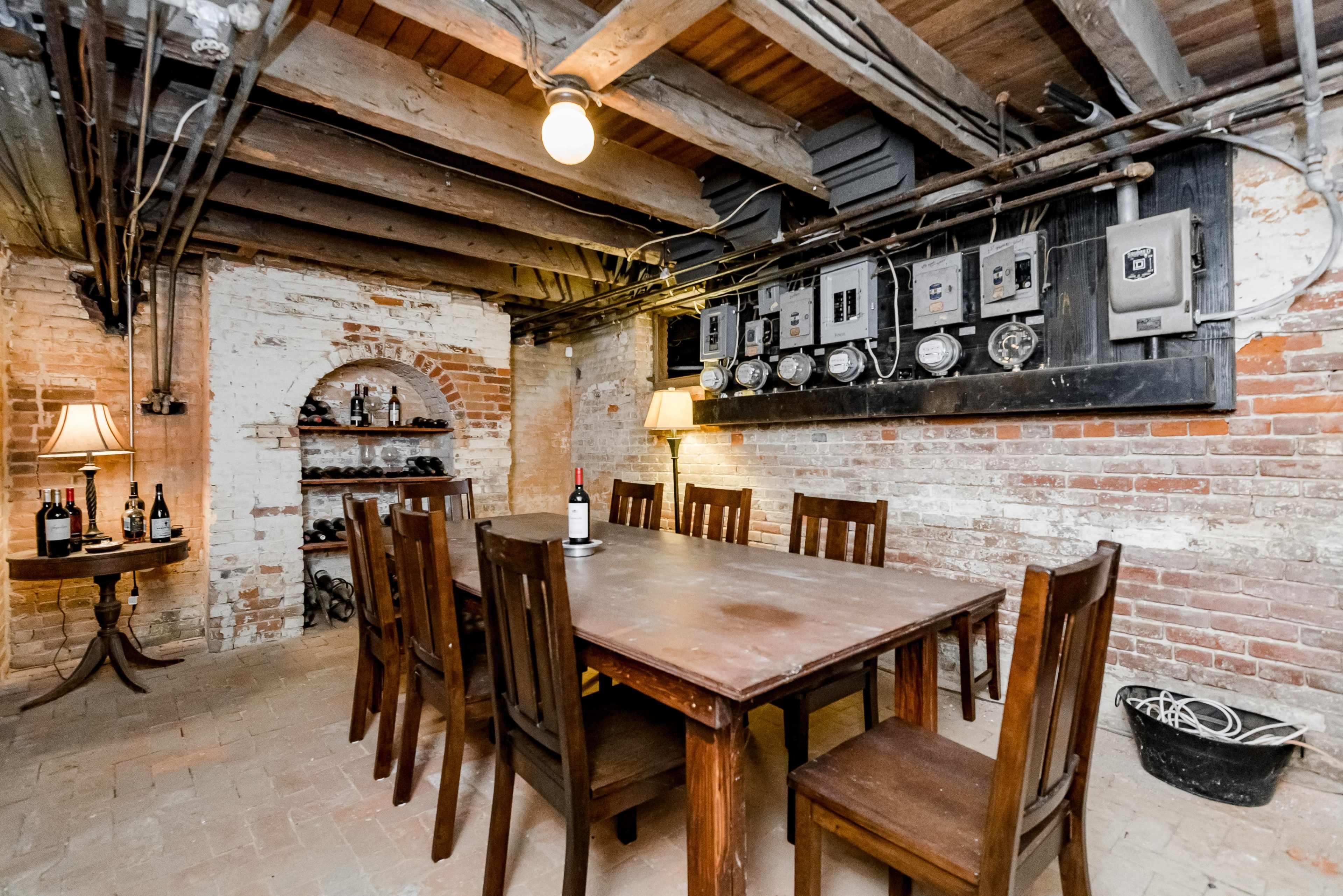 A rustic basement dining room with a large wooden table surrounded by chairs, flanked by exposed brick walls and overhead wooden beams.