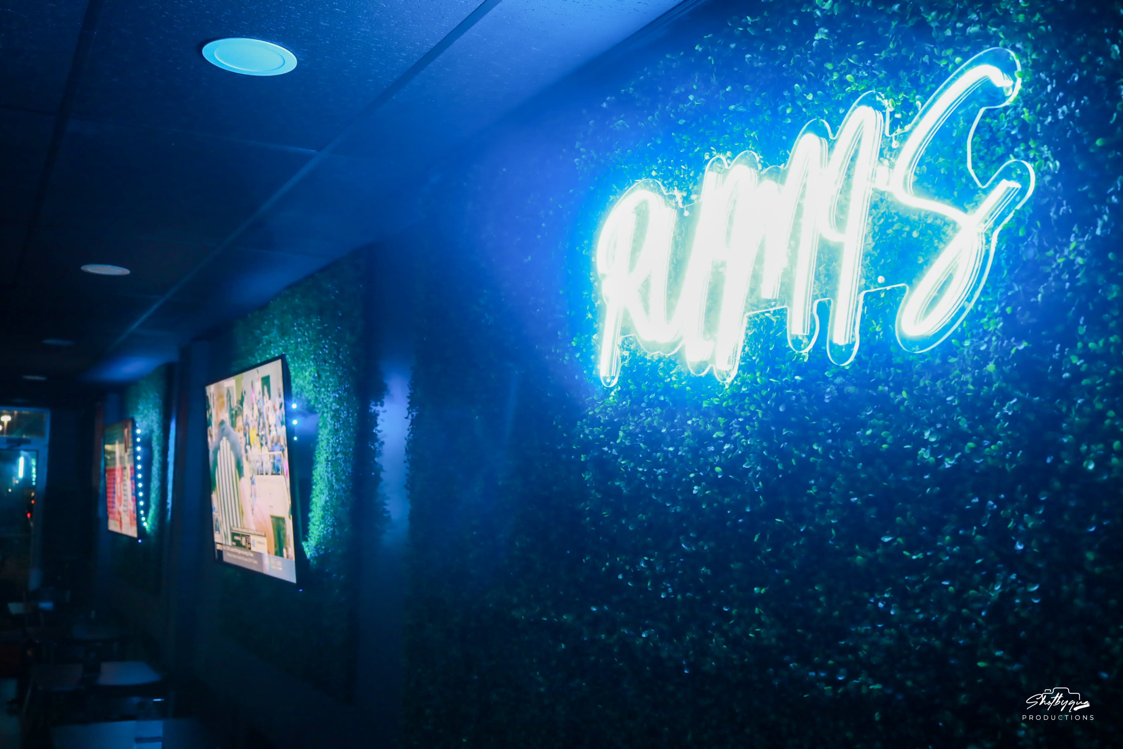 A neon sign reading "RUMS" is illuminated against a green wall with two screens displaying sports content.