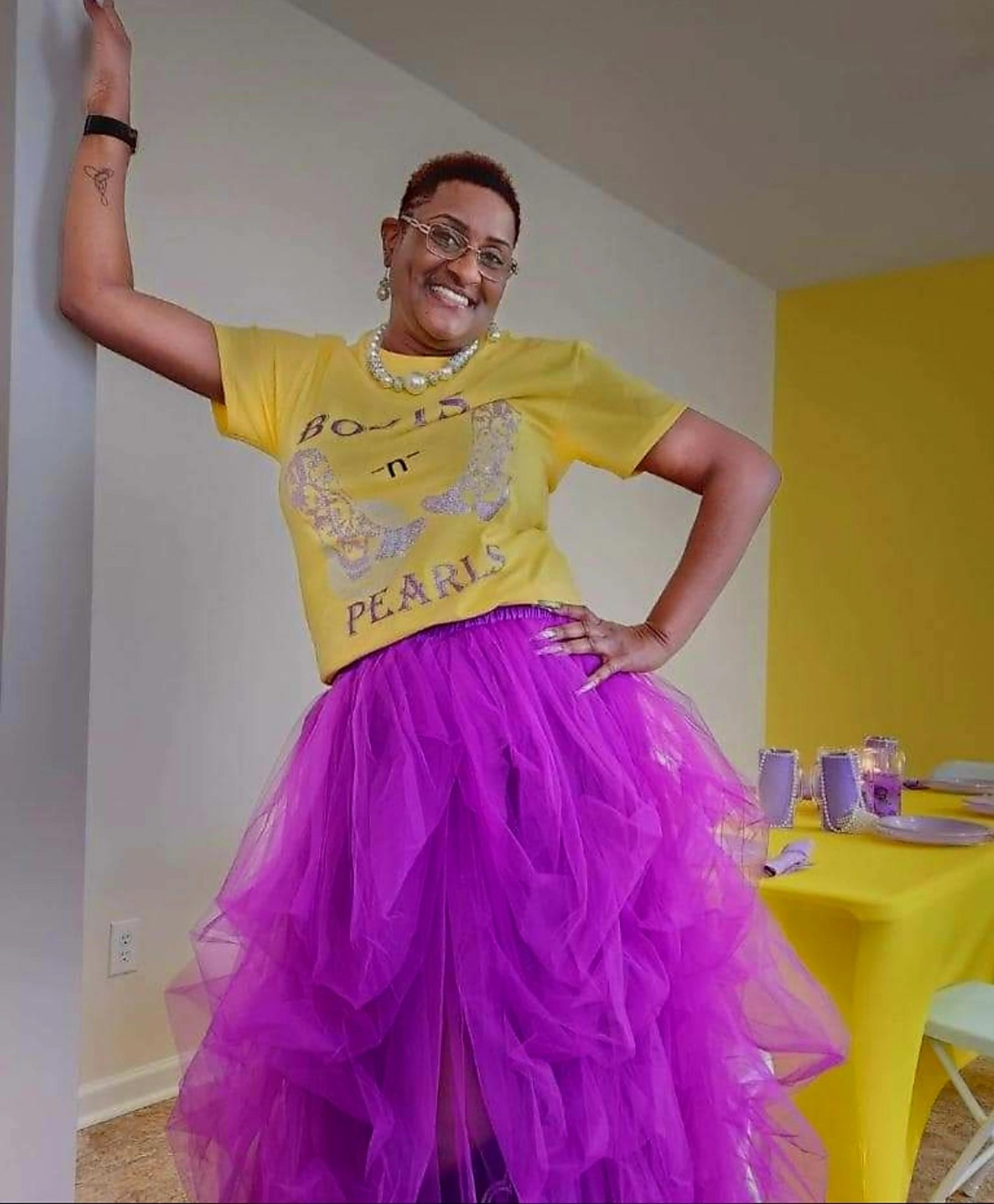 A woman stands in a brightly colored room, wearing a yellow graphic t-shirt and a purple tulle skirt, posing with one hand on her hip.