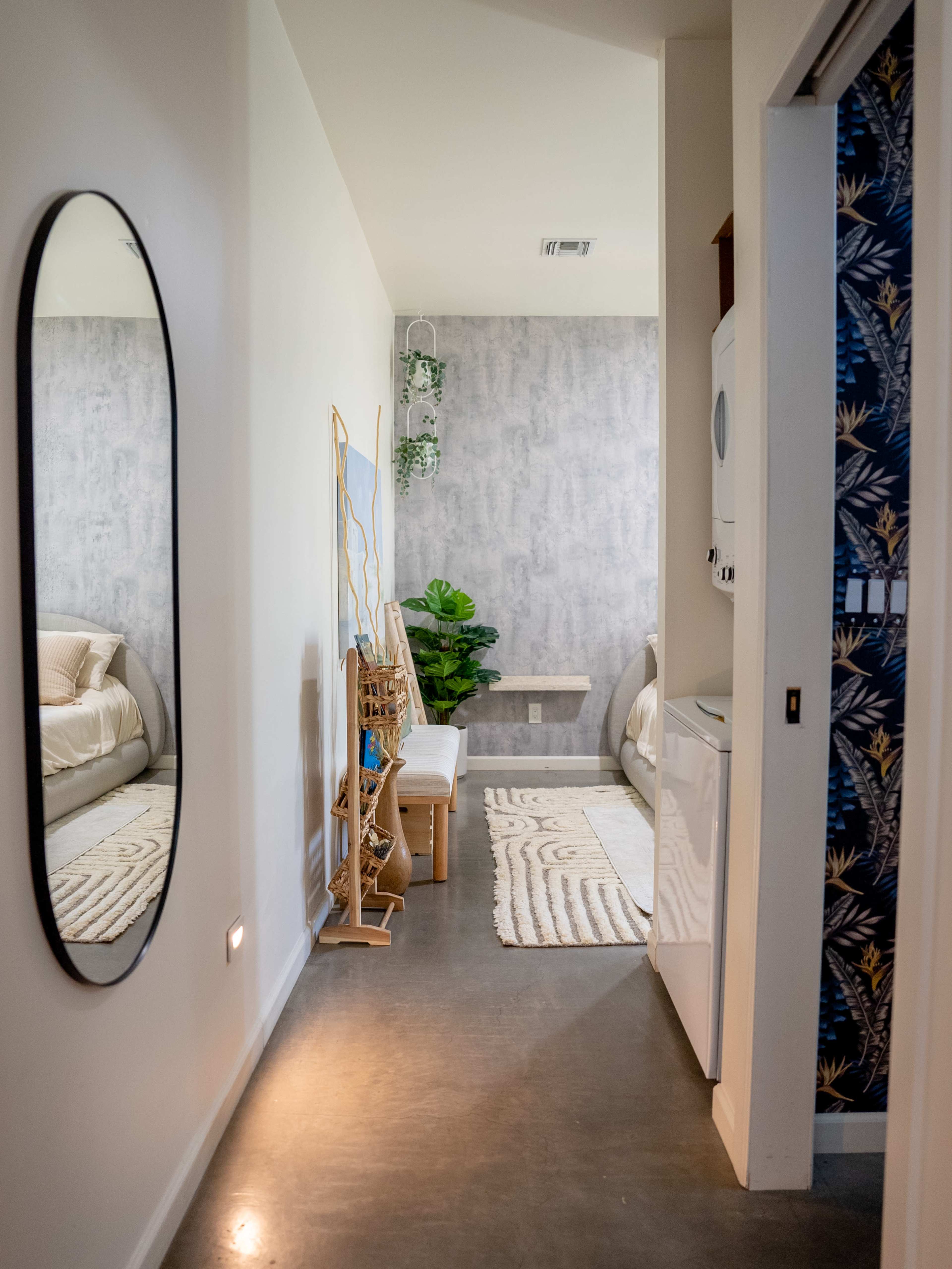 A narrow hallway leads to a bedroom, featuring a mirror, a plant, and a washing machine beside a decorative wall.