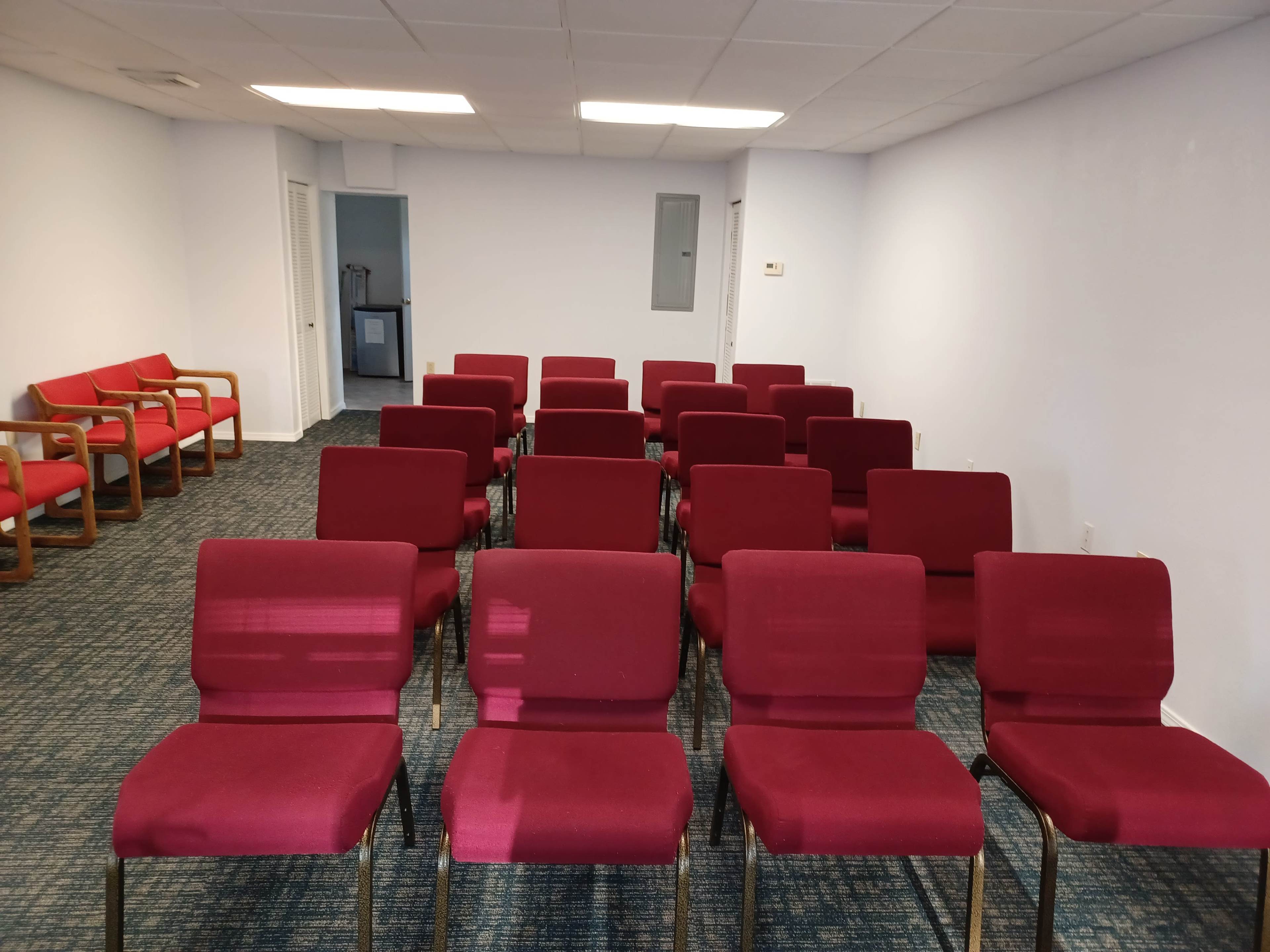 The image shows a room with rows of red chairs arranged in a grid pattern on a carpeted floor, with additional chairs positioned along one wall.