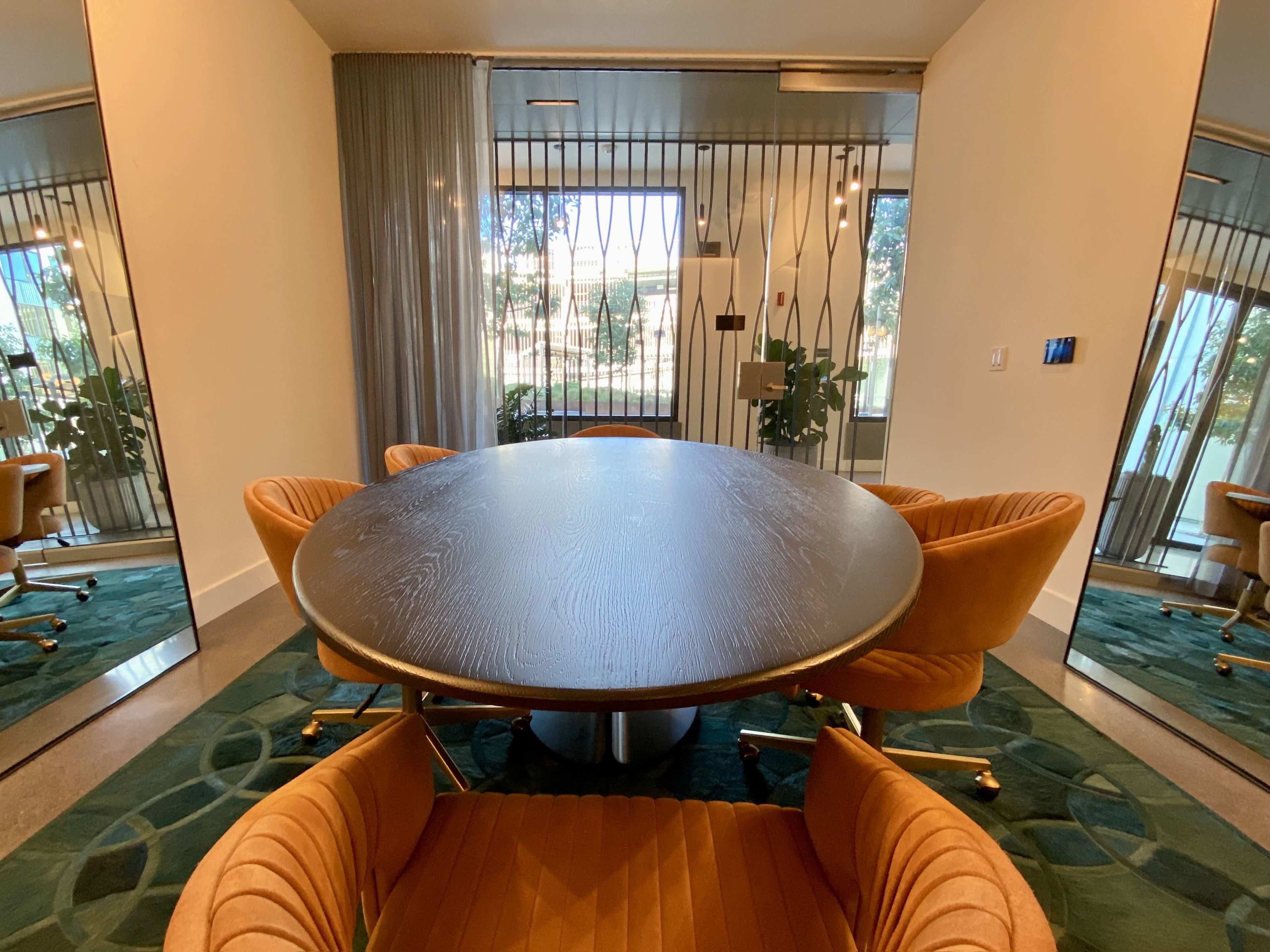 A round wooden table surrounded by four orange chairs is positioned in a room with large mirrors and glass doors leading to a garden.