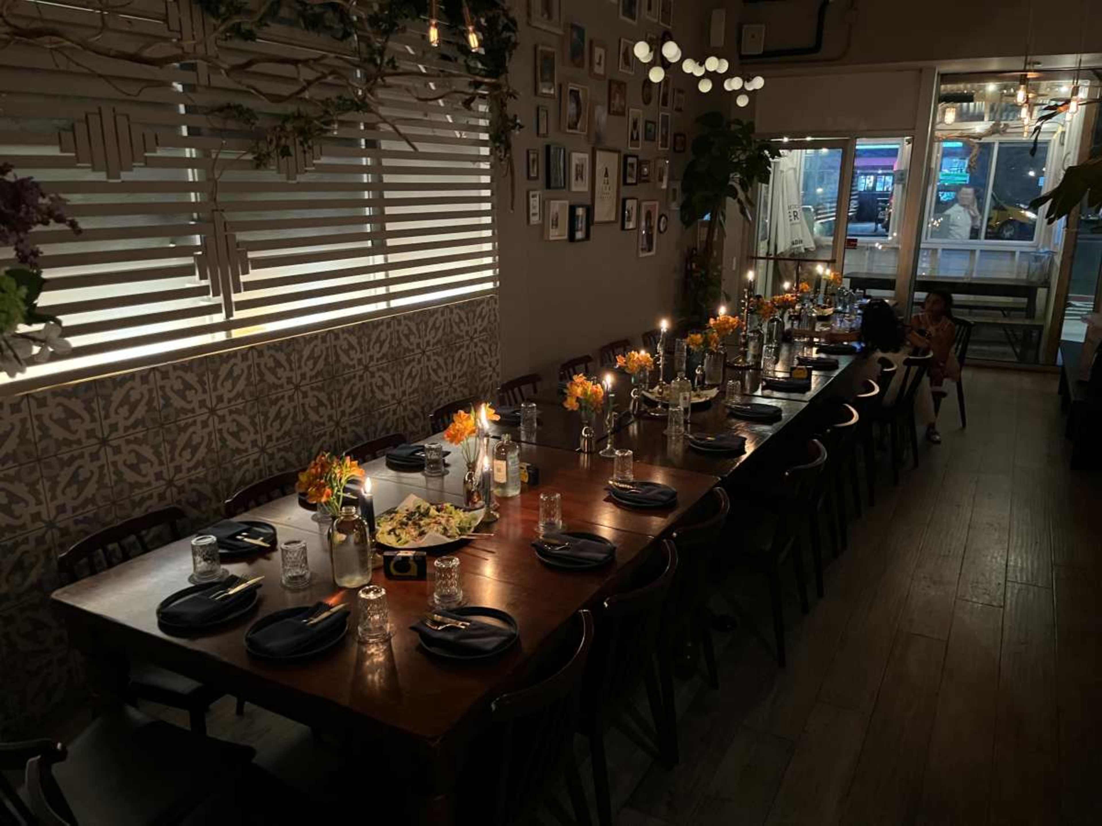 A long wooden table is set for dining, adorned with flowers and candles, in a softly lit room featuring framed pictures on the walls.