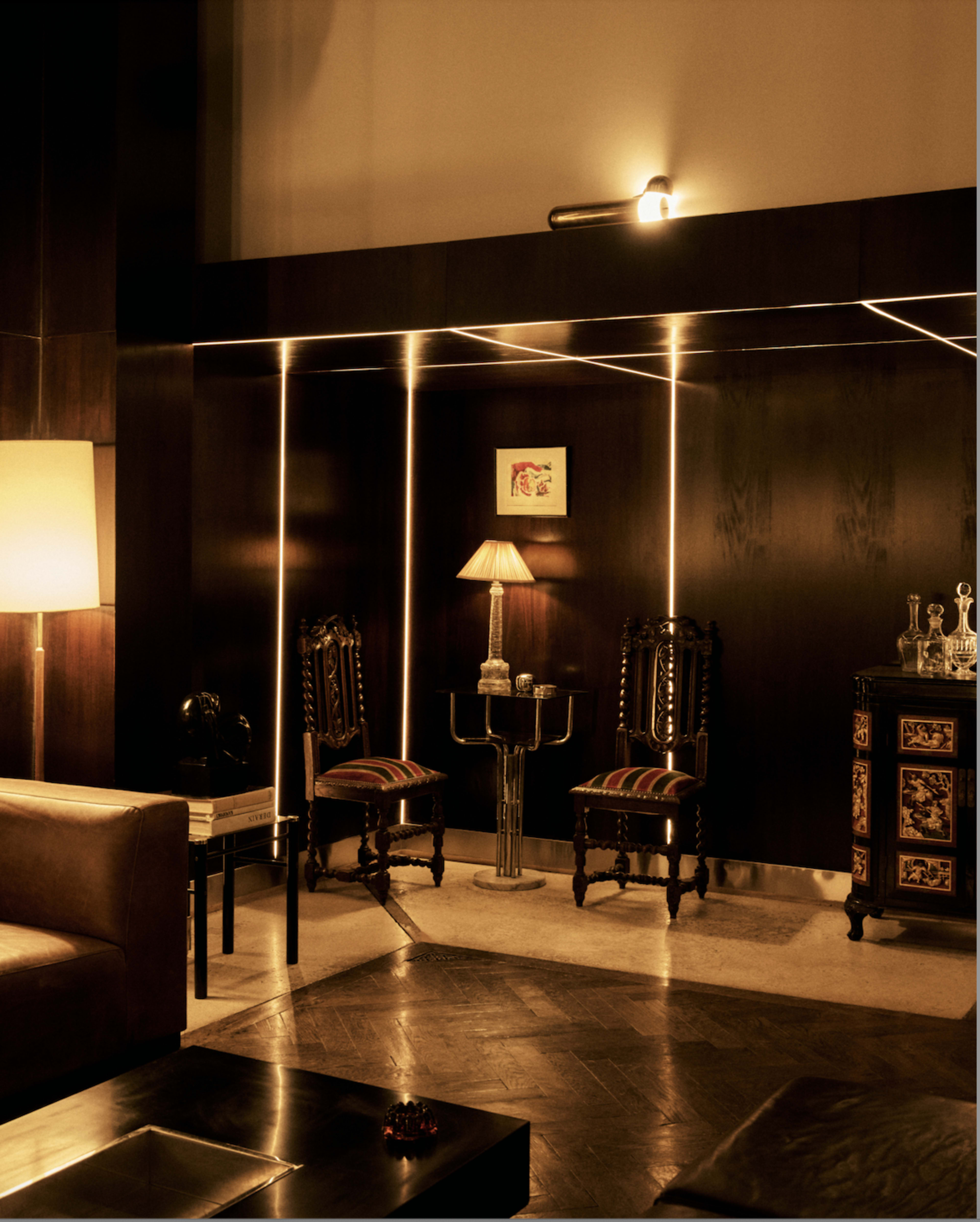 The image depicts a dimly lit lounge area with two ornate chairs, a lamp, a small table, and a vintage cabinet against a dark wall.
