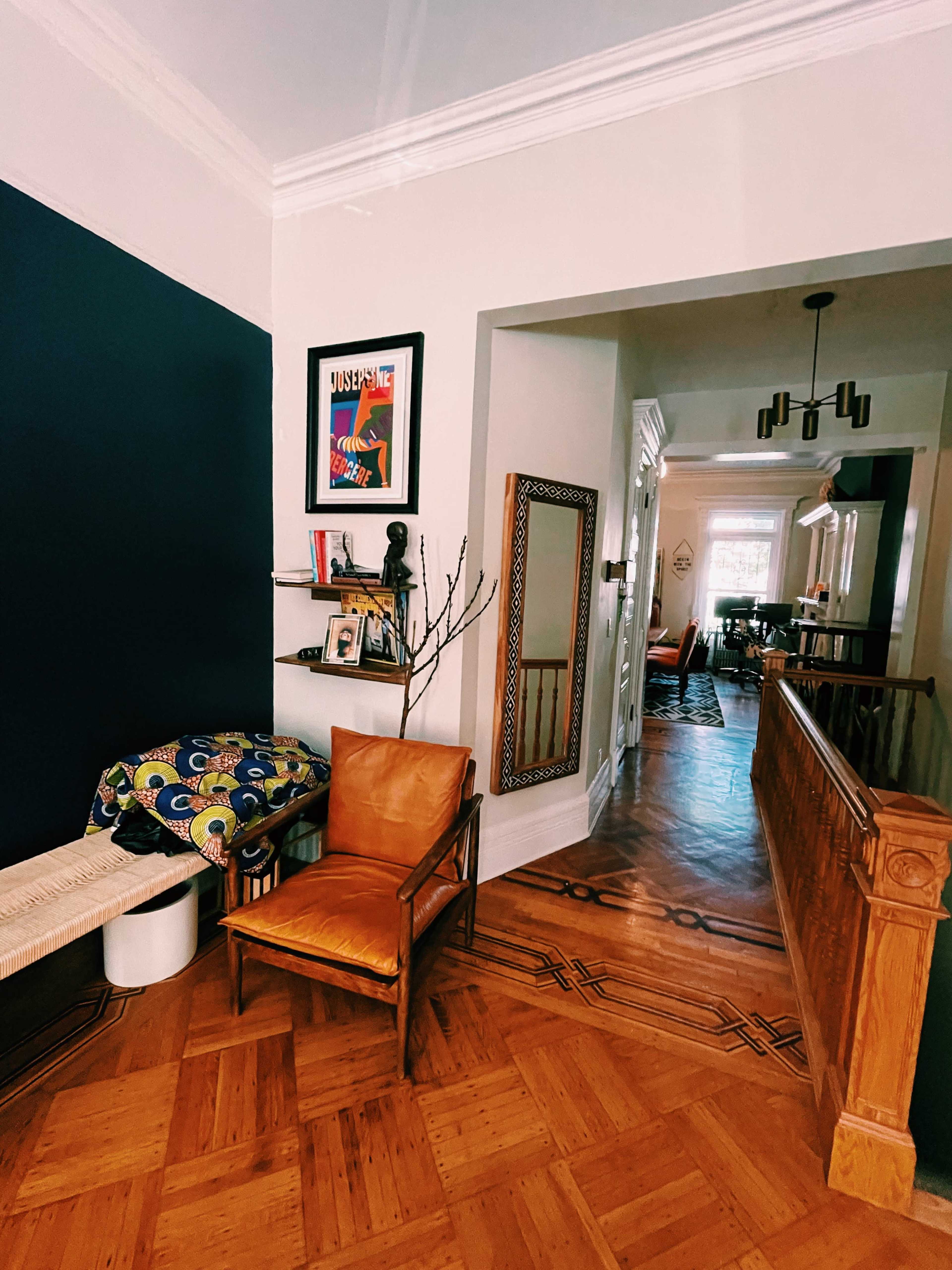 A hallway with a chair, a small shelf displaying framed pictures, and a table with decorative items, leading to another area of the home.