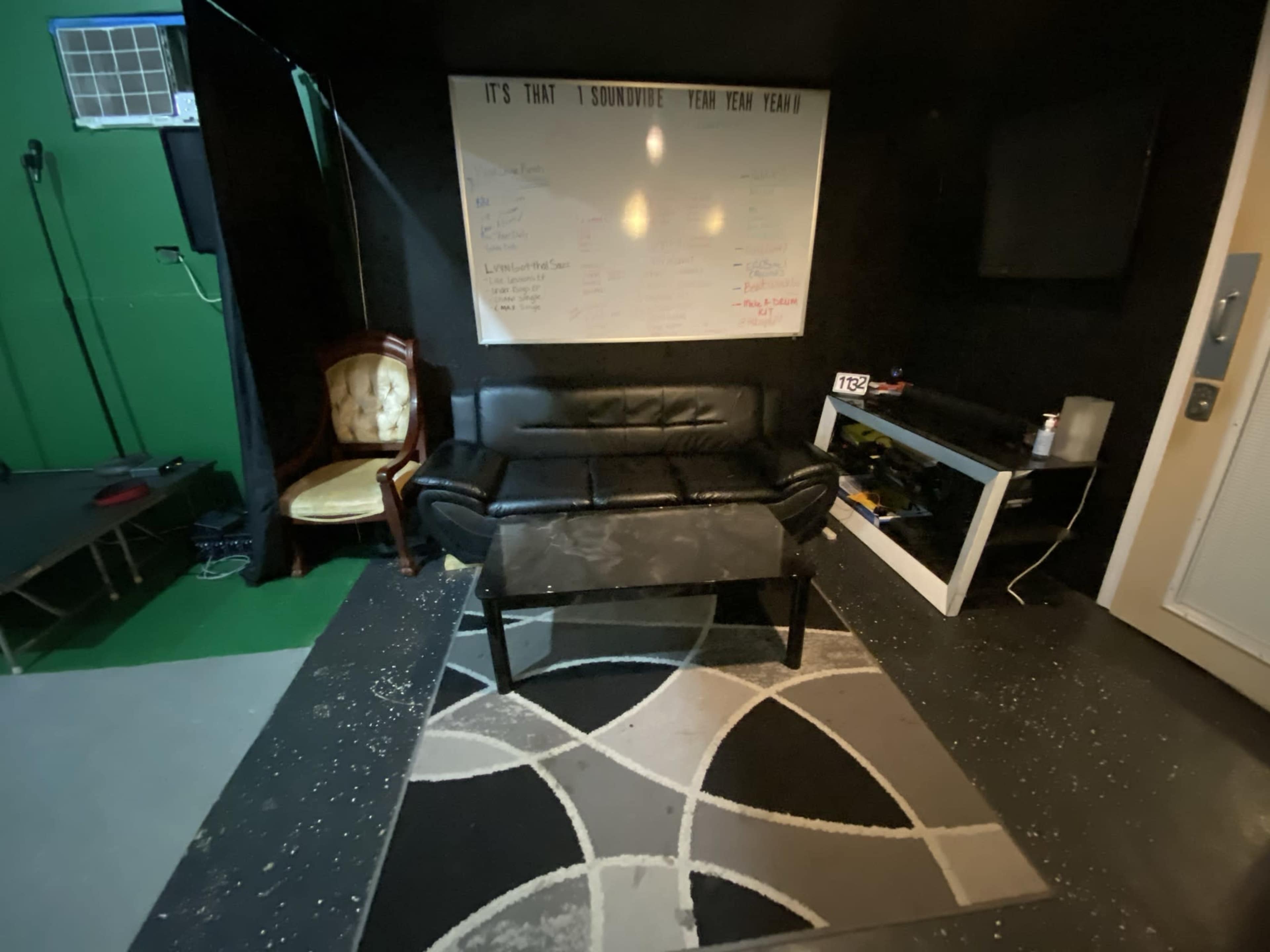 A black leather couch and a coffee table are positioned on a patterned rug in a dimly lit room with a whiteboard and television against one wall.