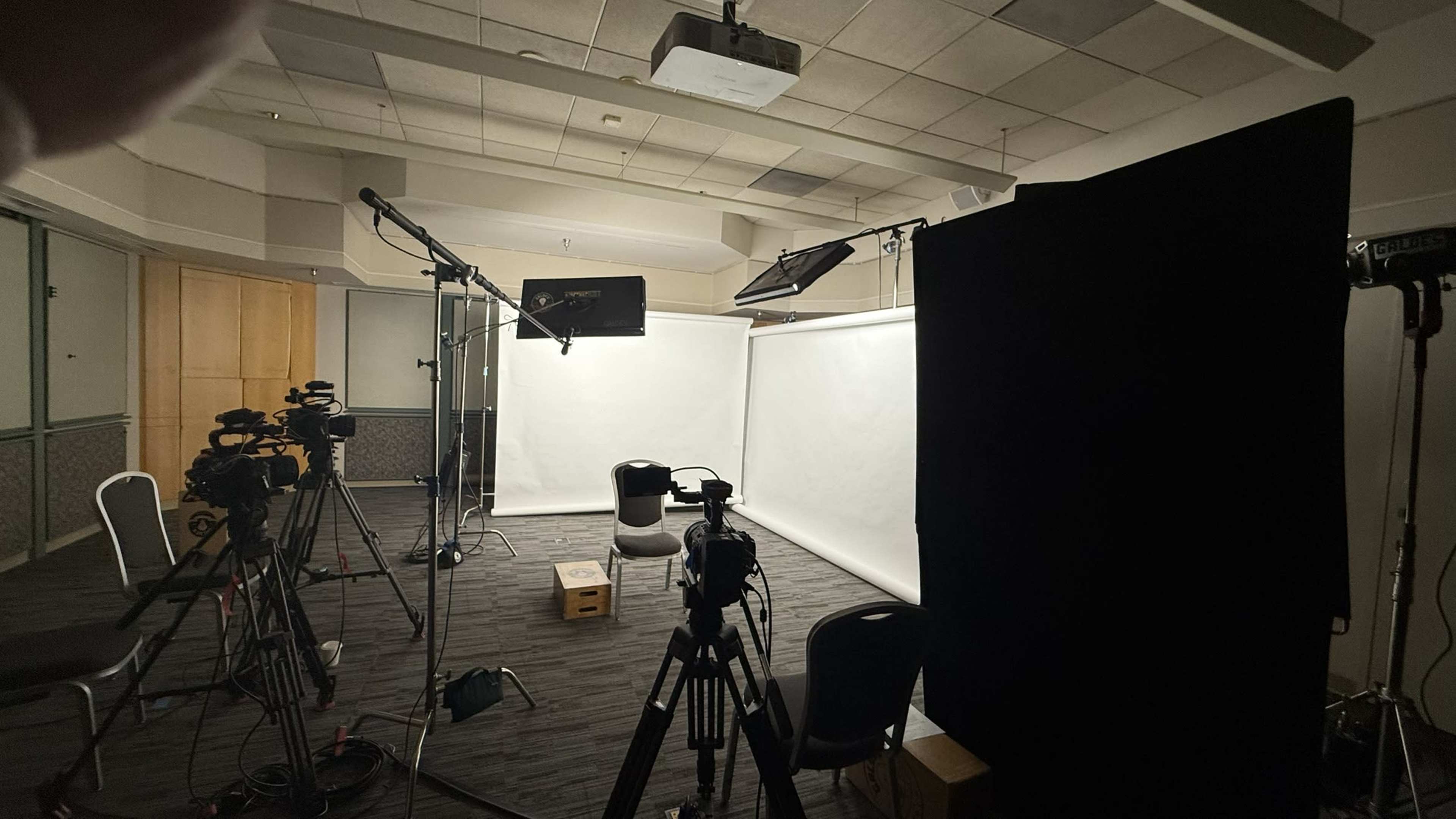 The image shows a dimly lit studio setup with multiple cameras, lighting equipment, and a black curtain in a space prepared for filming.