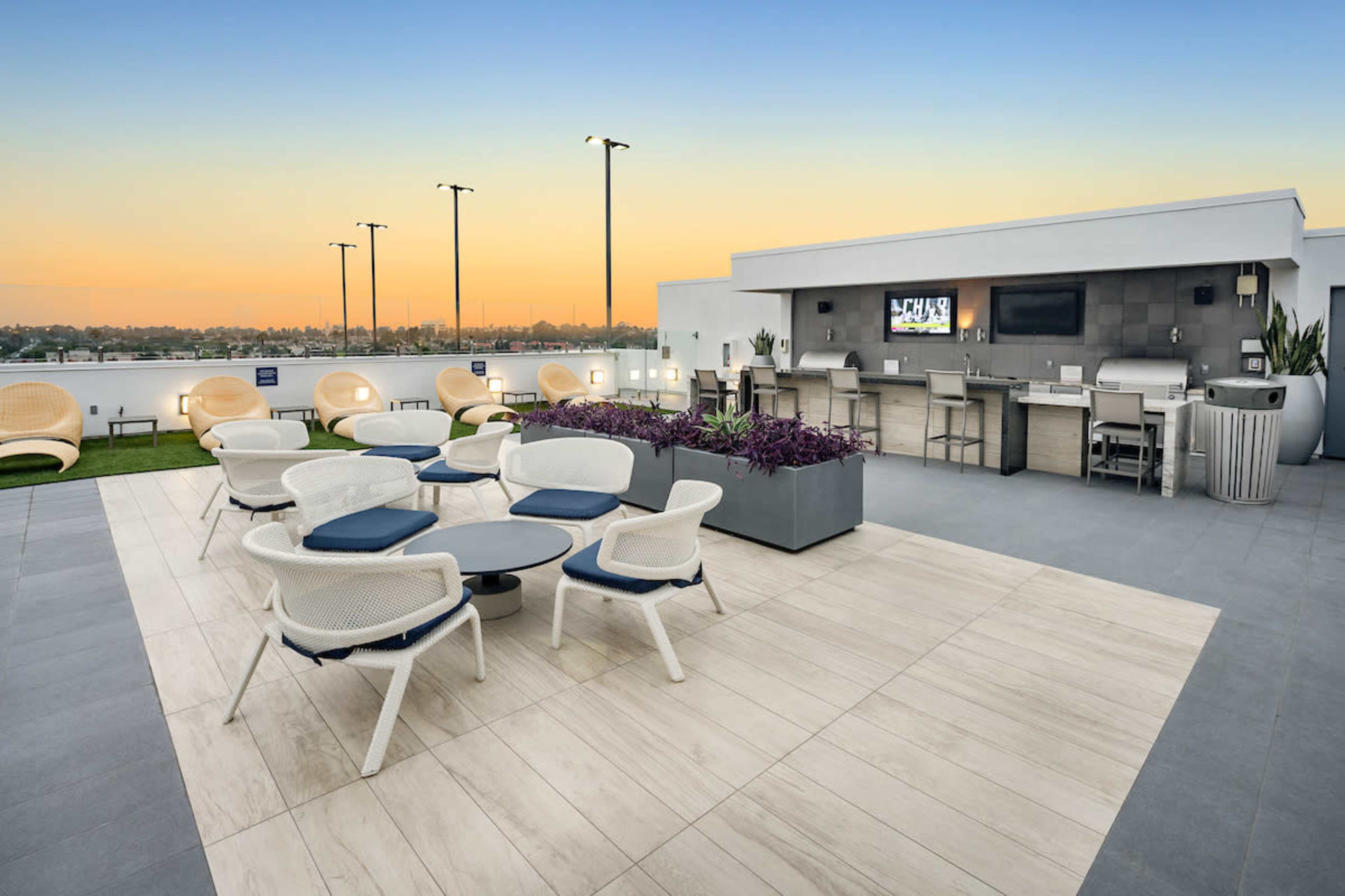 A rooftop terrace featuring modern seating arrangements, a bar area, and a view of the skyline at sunset.