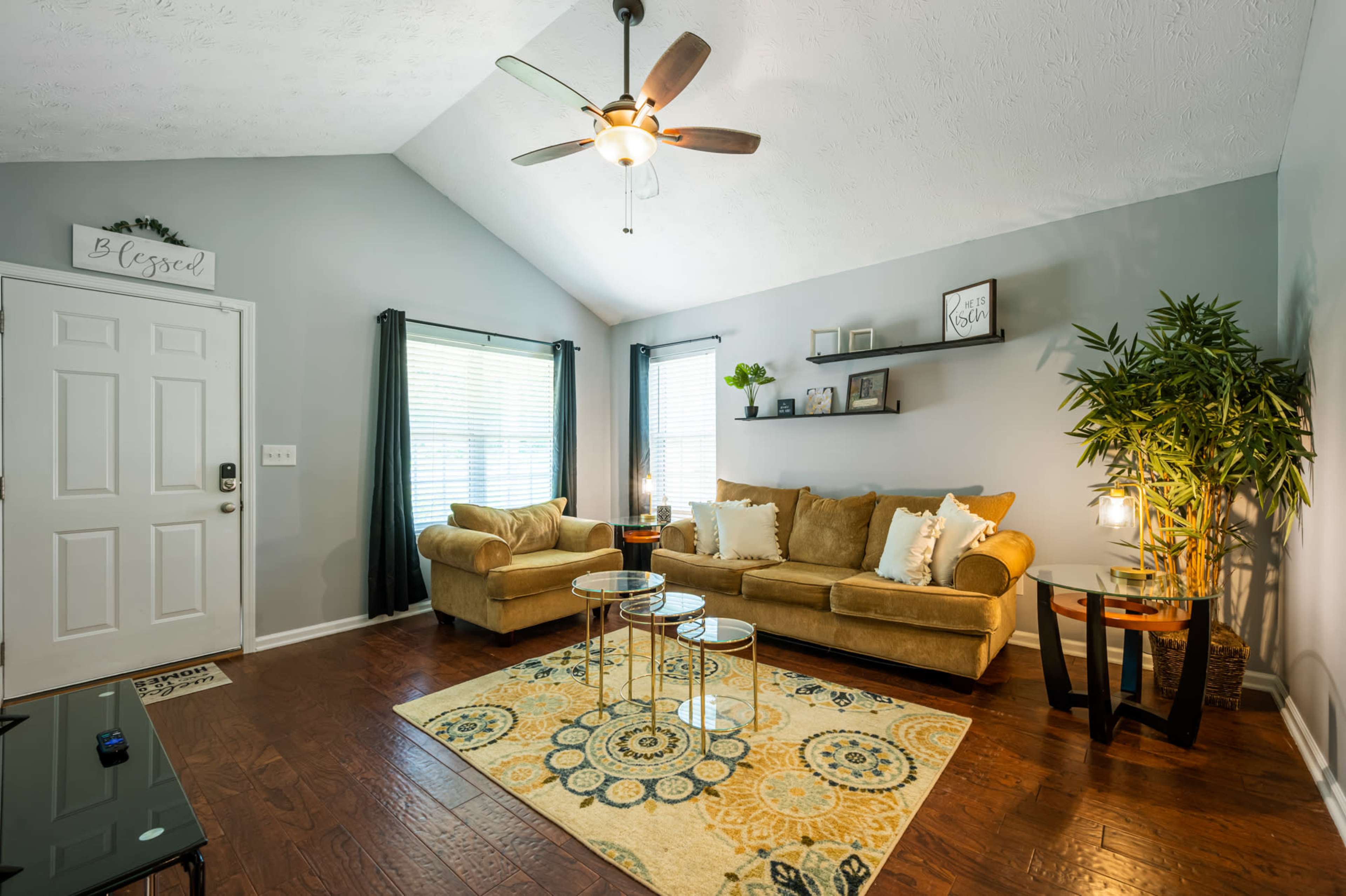 A living room features a beige couch, a decorative area rug, and a ceiling fan, with natural light coming from two windows.