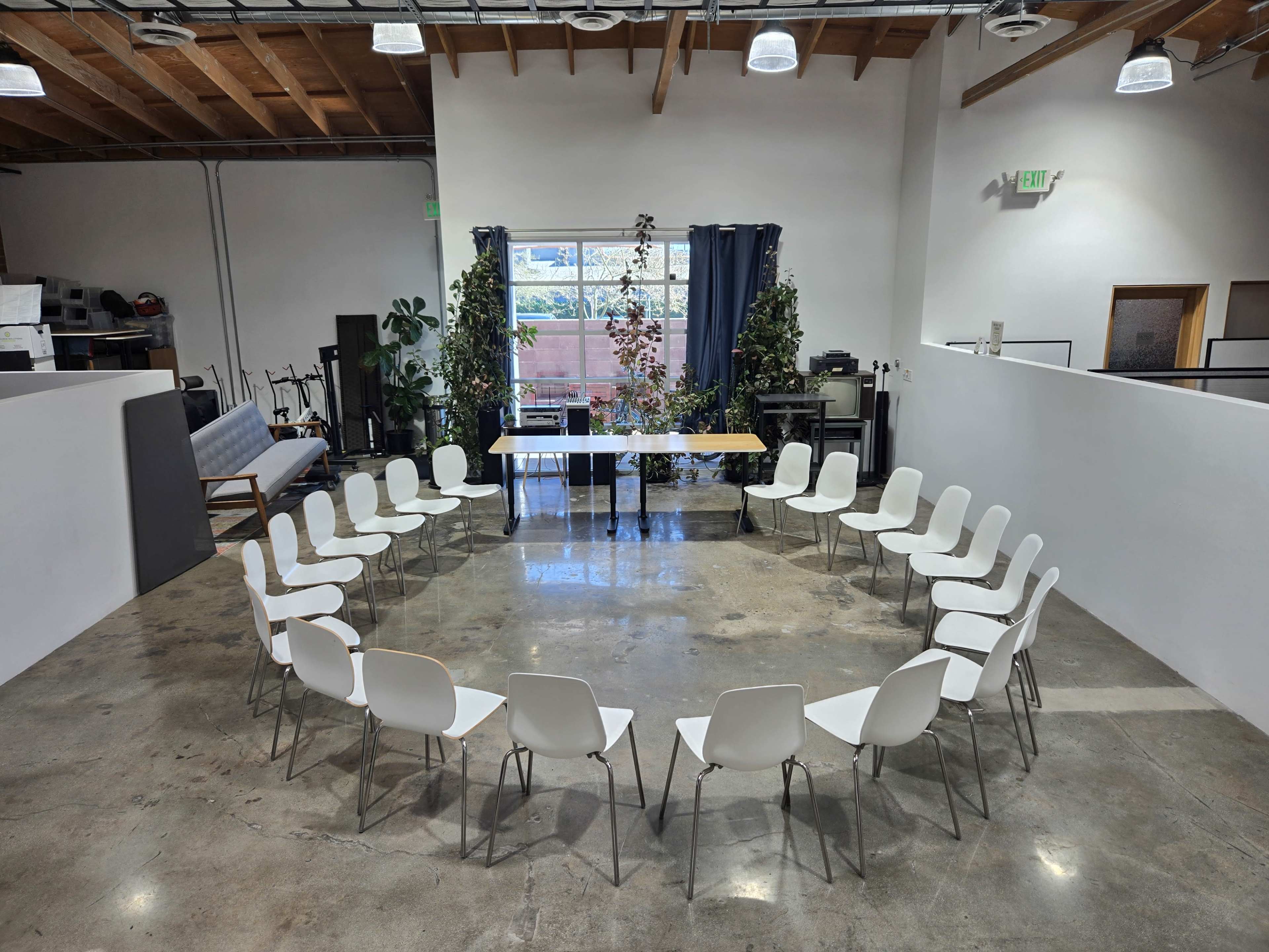 A circular arrangement of white chairs surrounds a long wooden table in a spacious, well-lit room with large windows and plants.