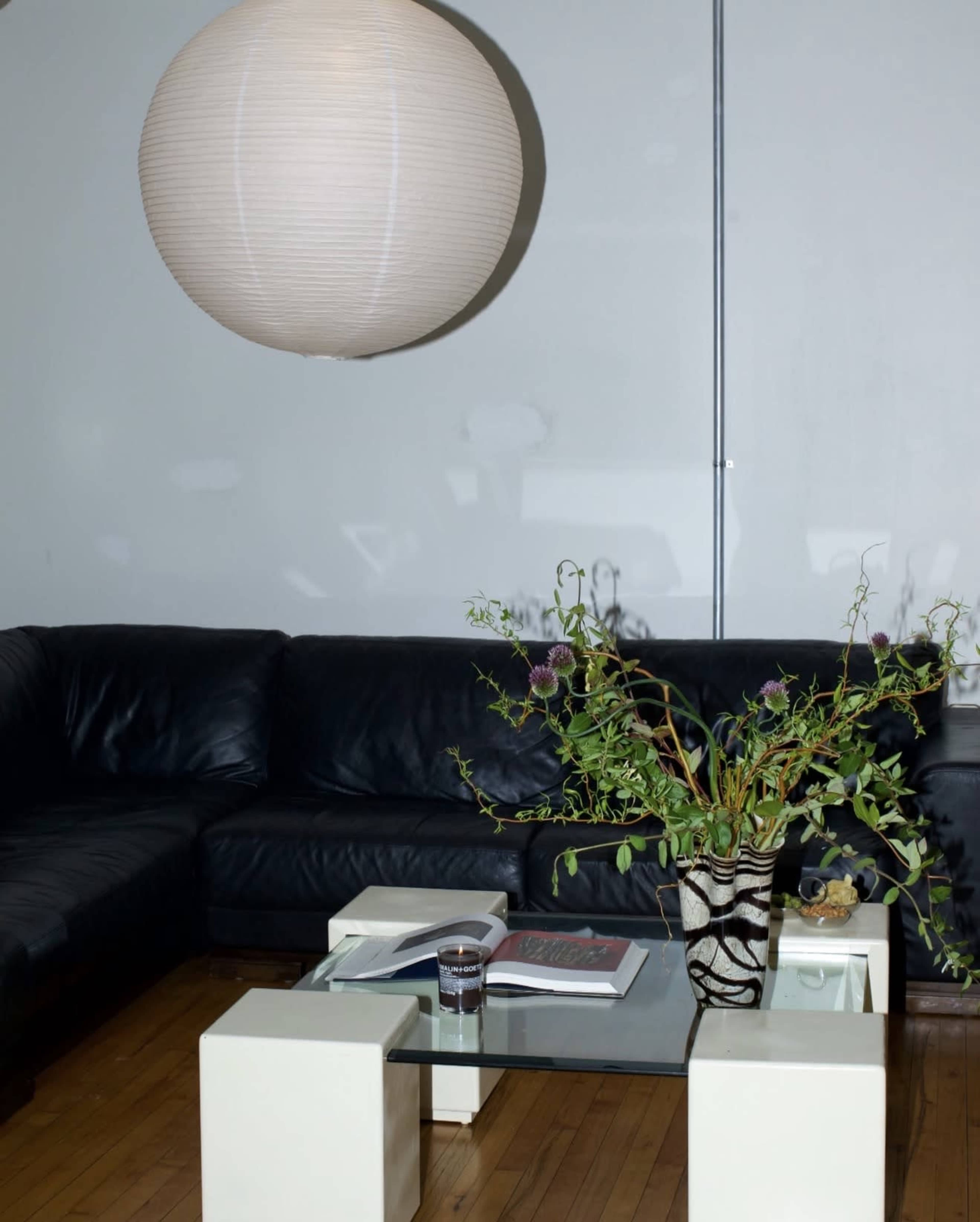 A modern living room features a black leather sectional sofa, a glass coffee table with a vase of plants, and a large paper lantern hanging above.