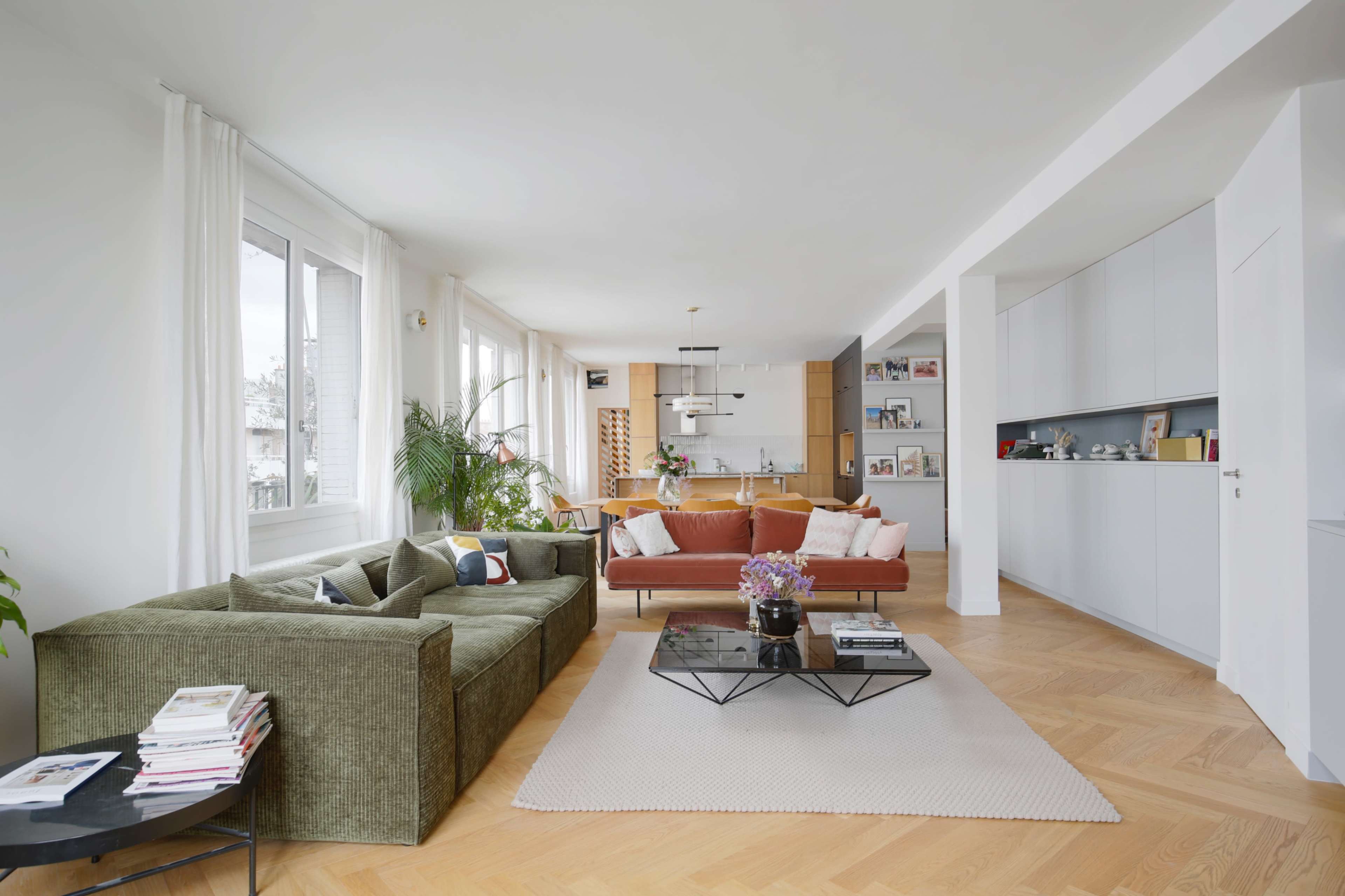 A modern living room featuring a green sofa, a brown couch, a central coffee table, and light wooden floors, with large windows providing natural light.
