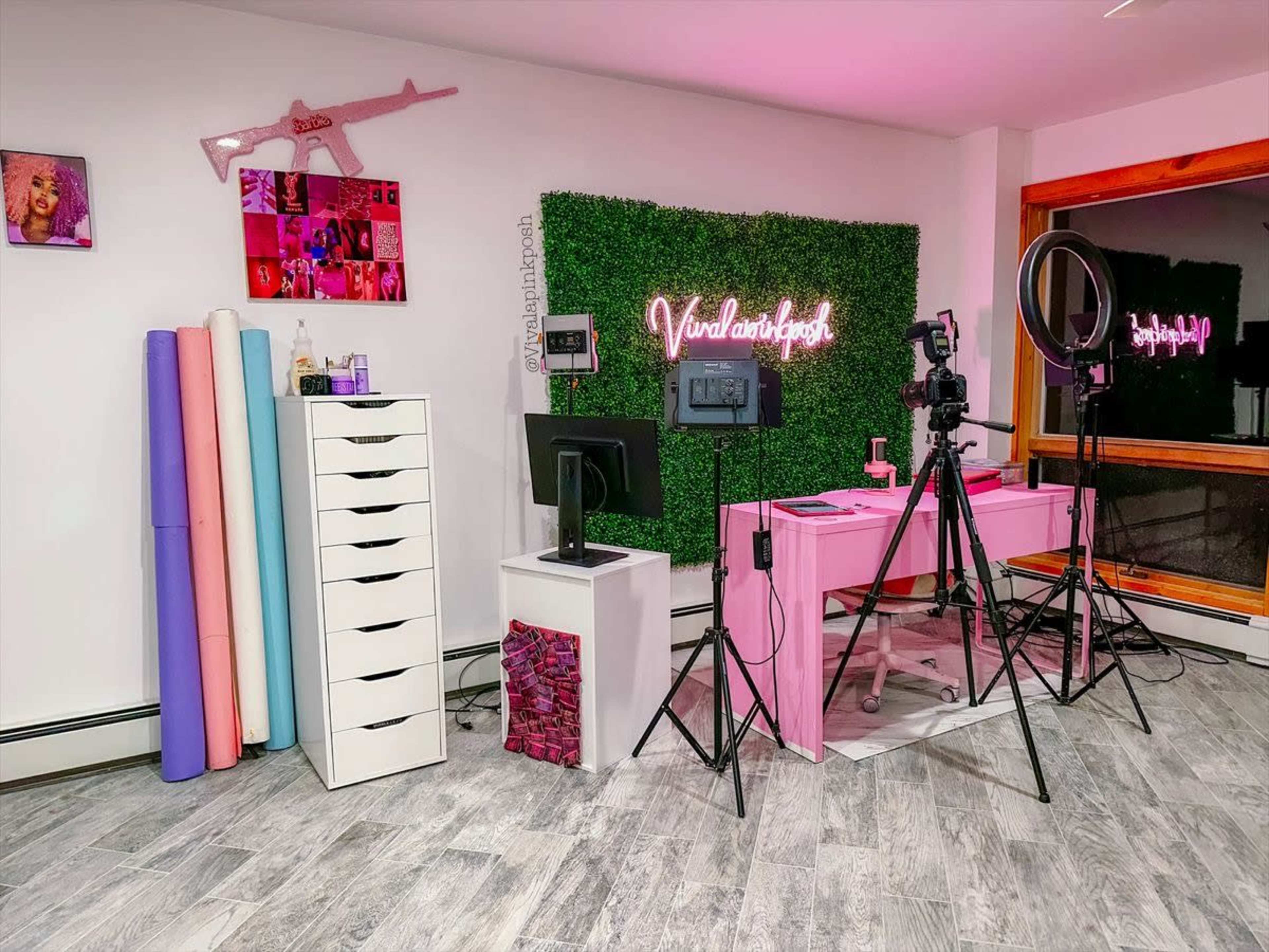 The image shows a brightly lit workspace featuring a pink desk, a black monitor, camera equipment on a tripod, and a variety of colorful backdrop rolls beside a white storage drawer unit.