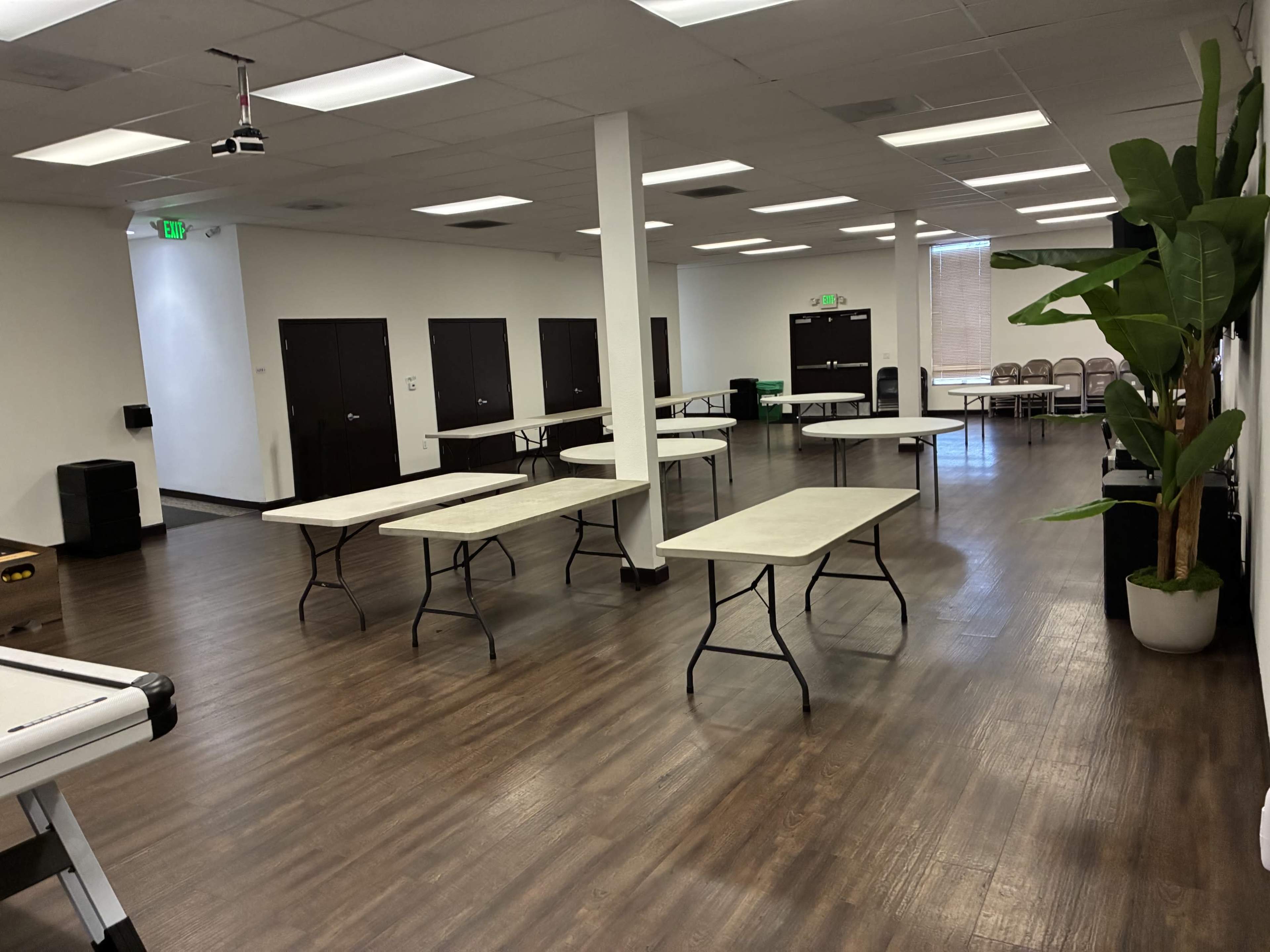 The image shows an empty room with several round tables arranged in rows, dark doors along one wall, and a large potted plant in the corner.