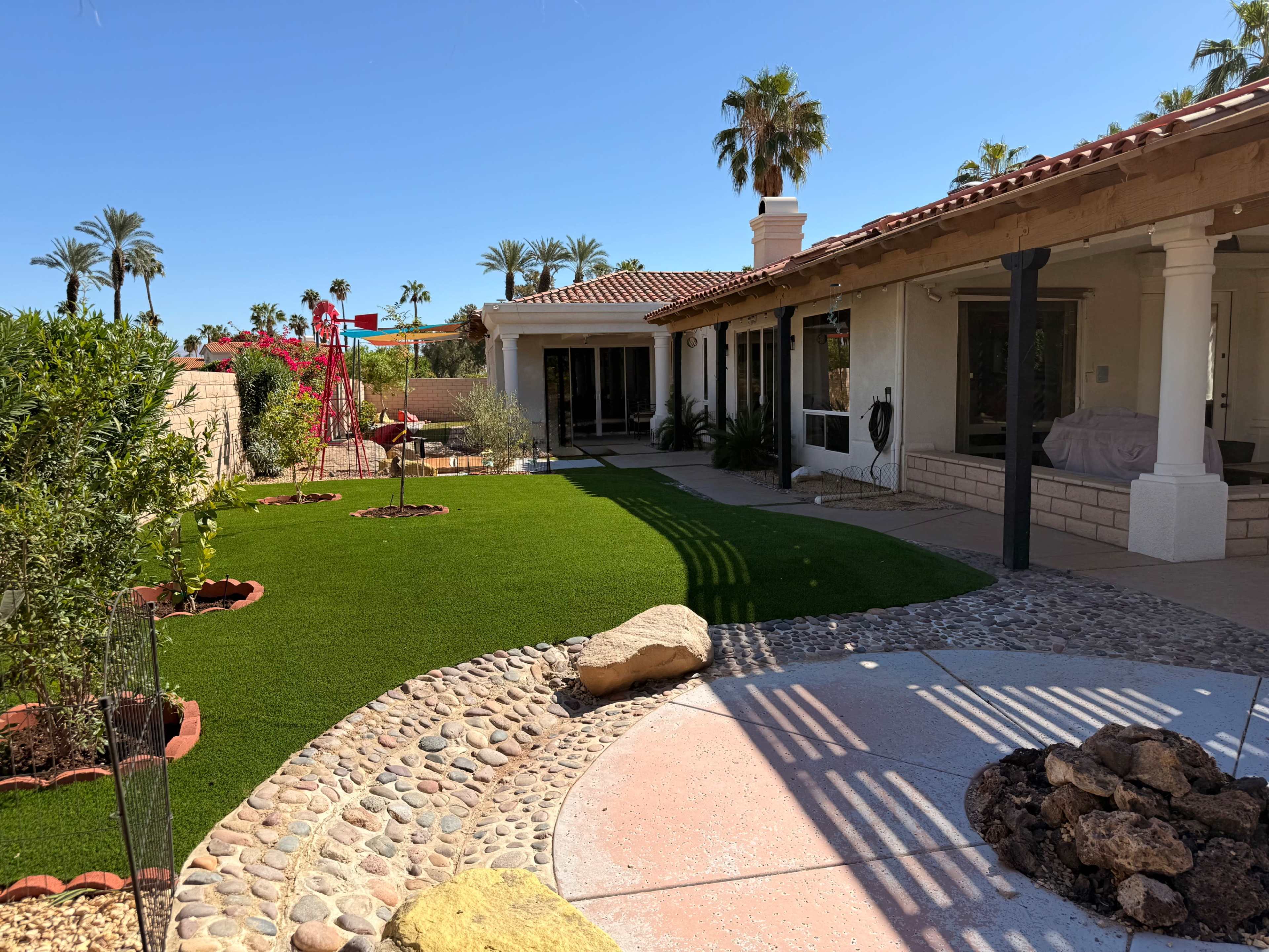 The image depicts a landscaped backyard featuring artificial turf, decorative stones, and a play structure, with a house and palm trees in the background.