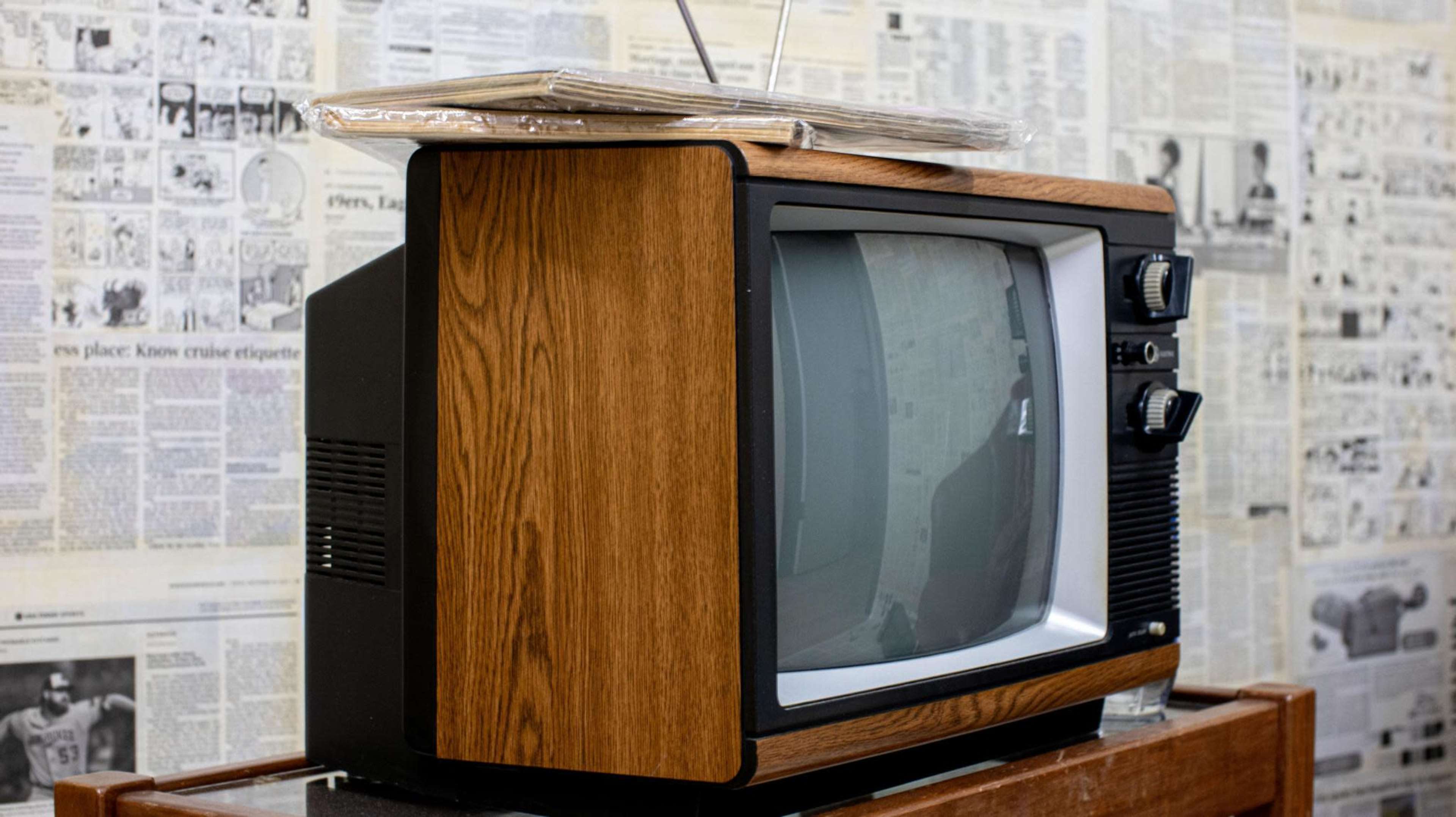 An old wooden television set with antennas sits on a table, surrounded by newspaper clippings on the wall.
