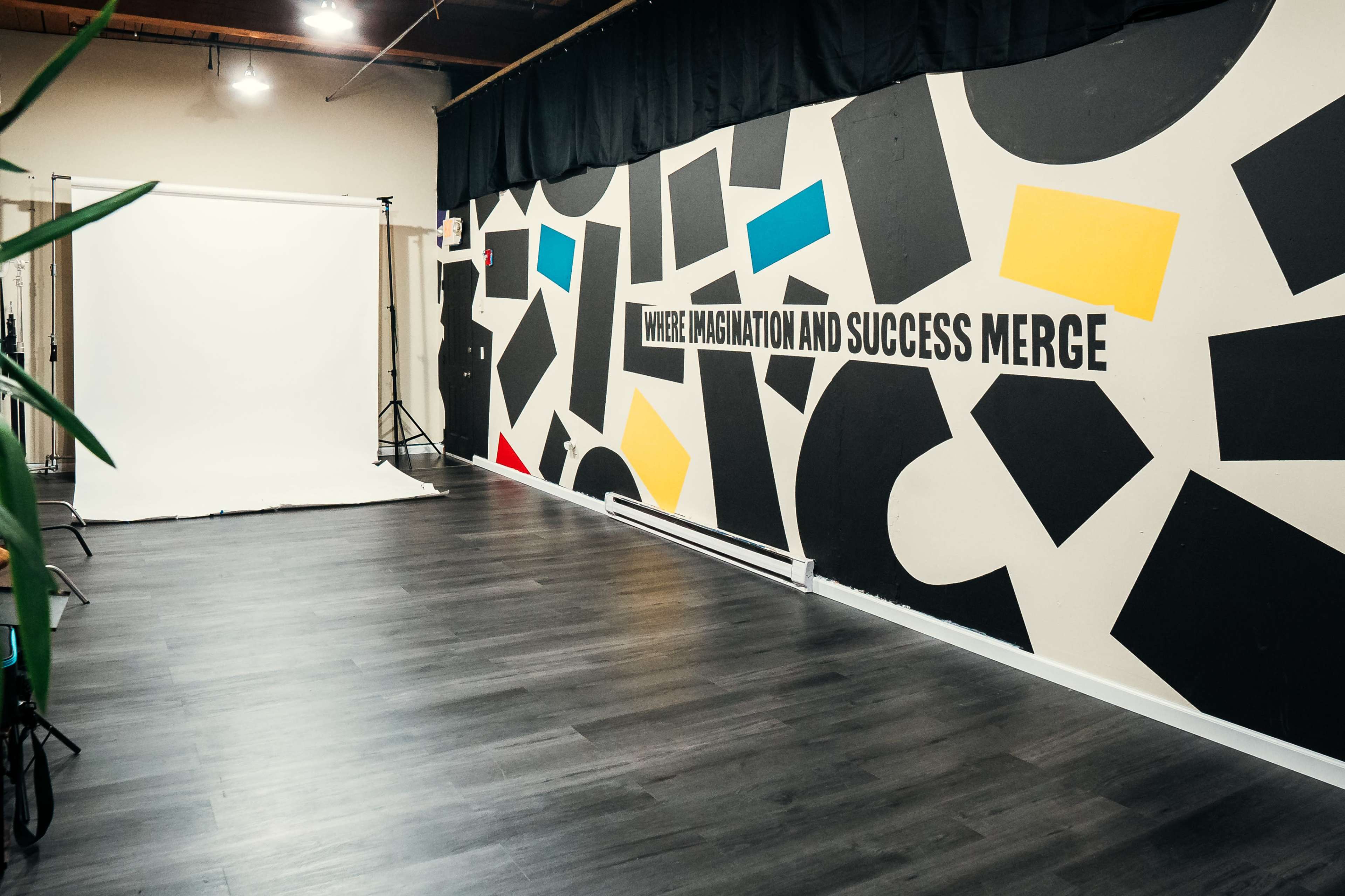 The image shows an empty room with a large wall featuring a graphic mural in black, yellow, blue, and red shapes, alongside a white backdrop set up for photography.