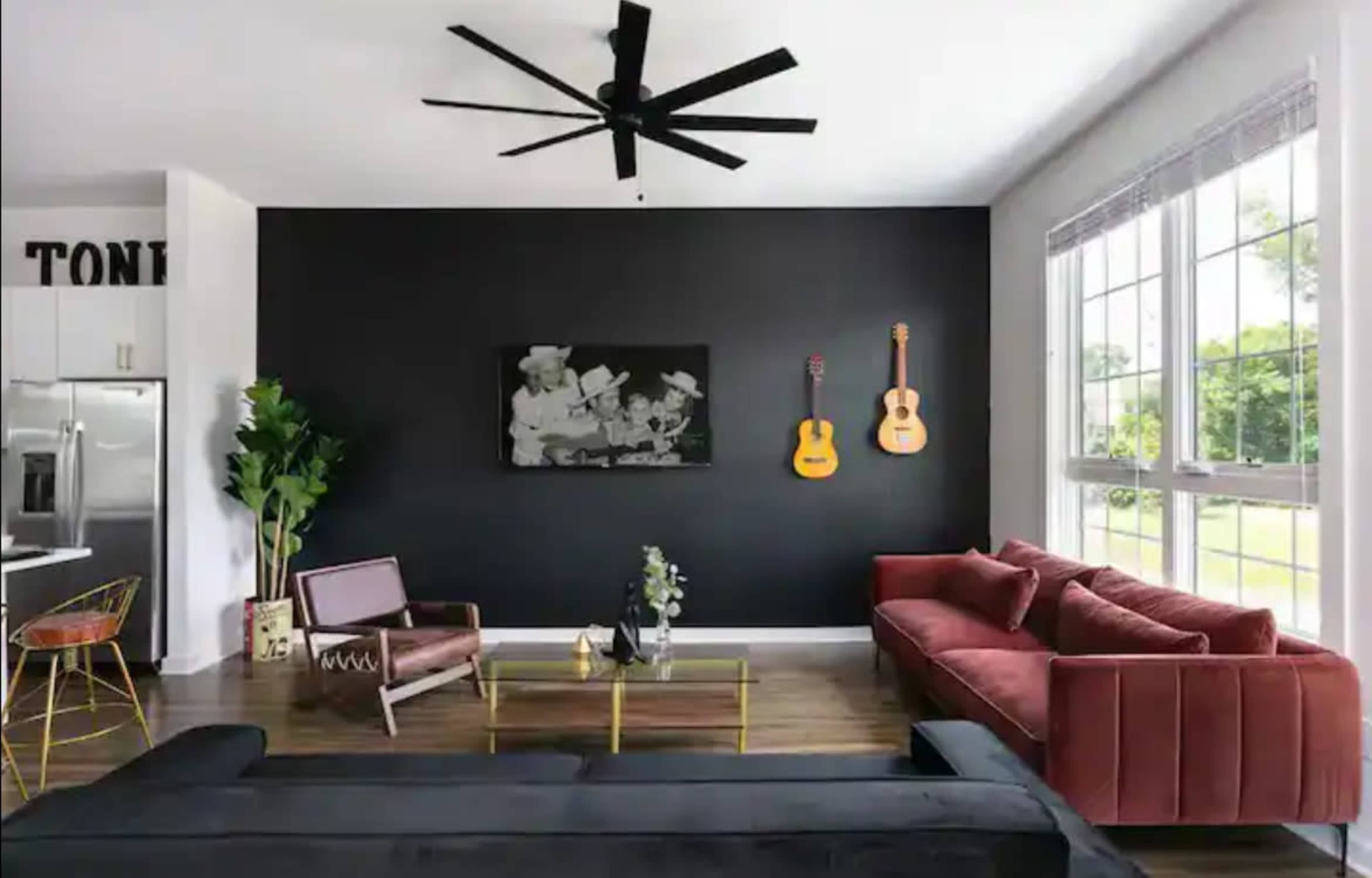 A modern living room features a dark accent wall with two acoustic guitars, a red sofa, a black sofa, a wooden coffee table, and large windows allowing natural light.
