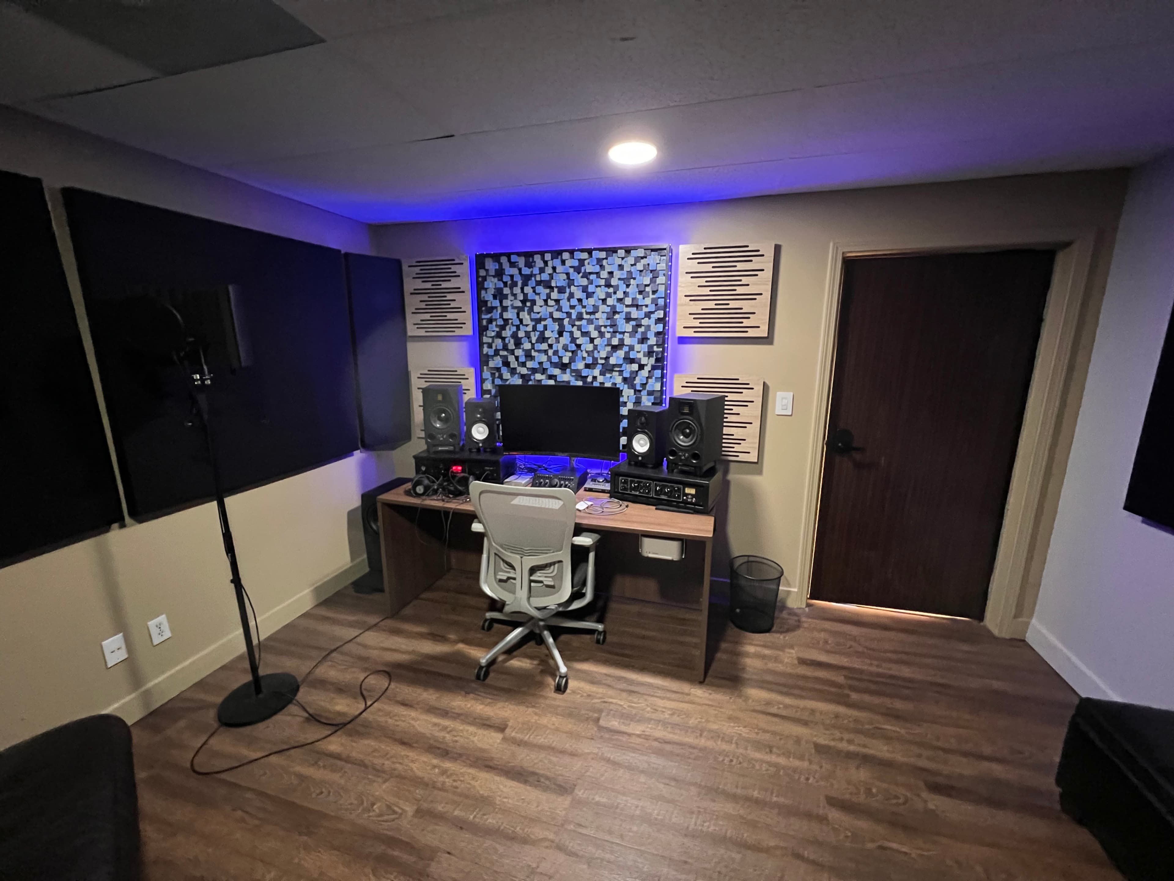 The image shows a music studio setup with a desk, computer, sound monitors, and acoustic panels on the walls, illuminated by blue LED lighting.