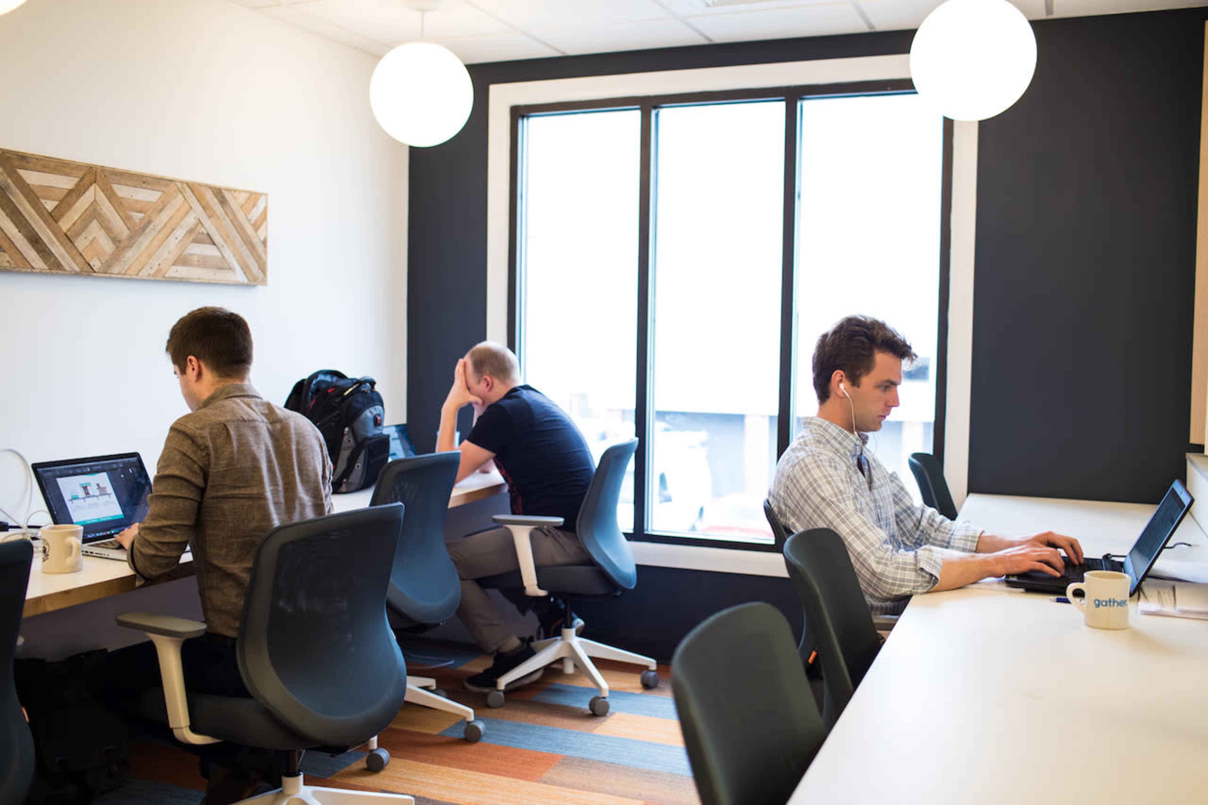 Three people are working on laptops at a shared office space with large windows and modern desks.
