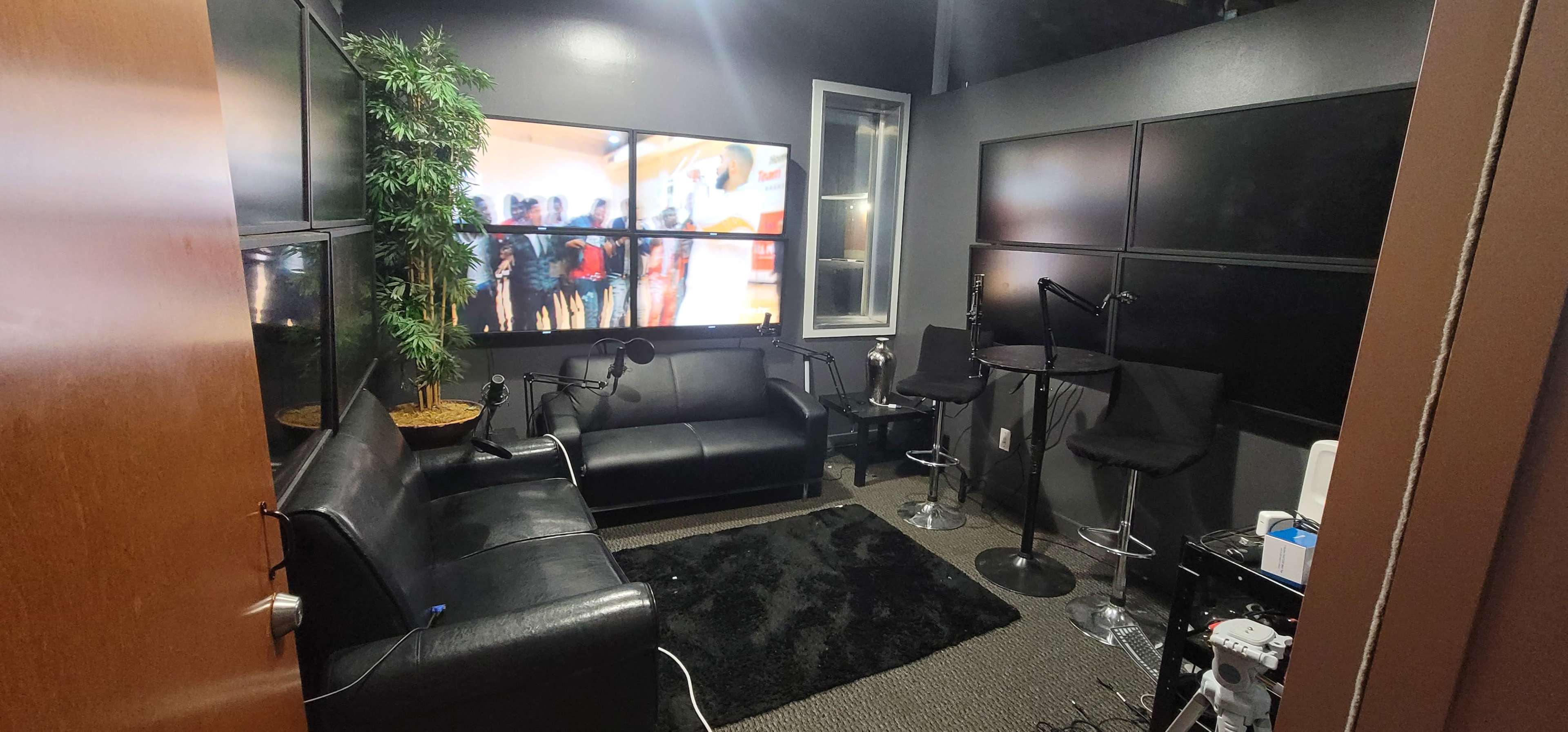 A small, modern room furnished with a black leather sofa, bar stools, and a large screen displaying people in another setting.