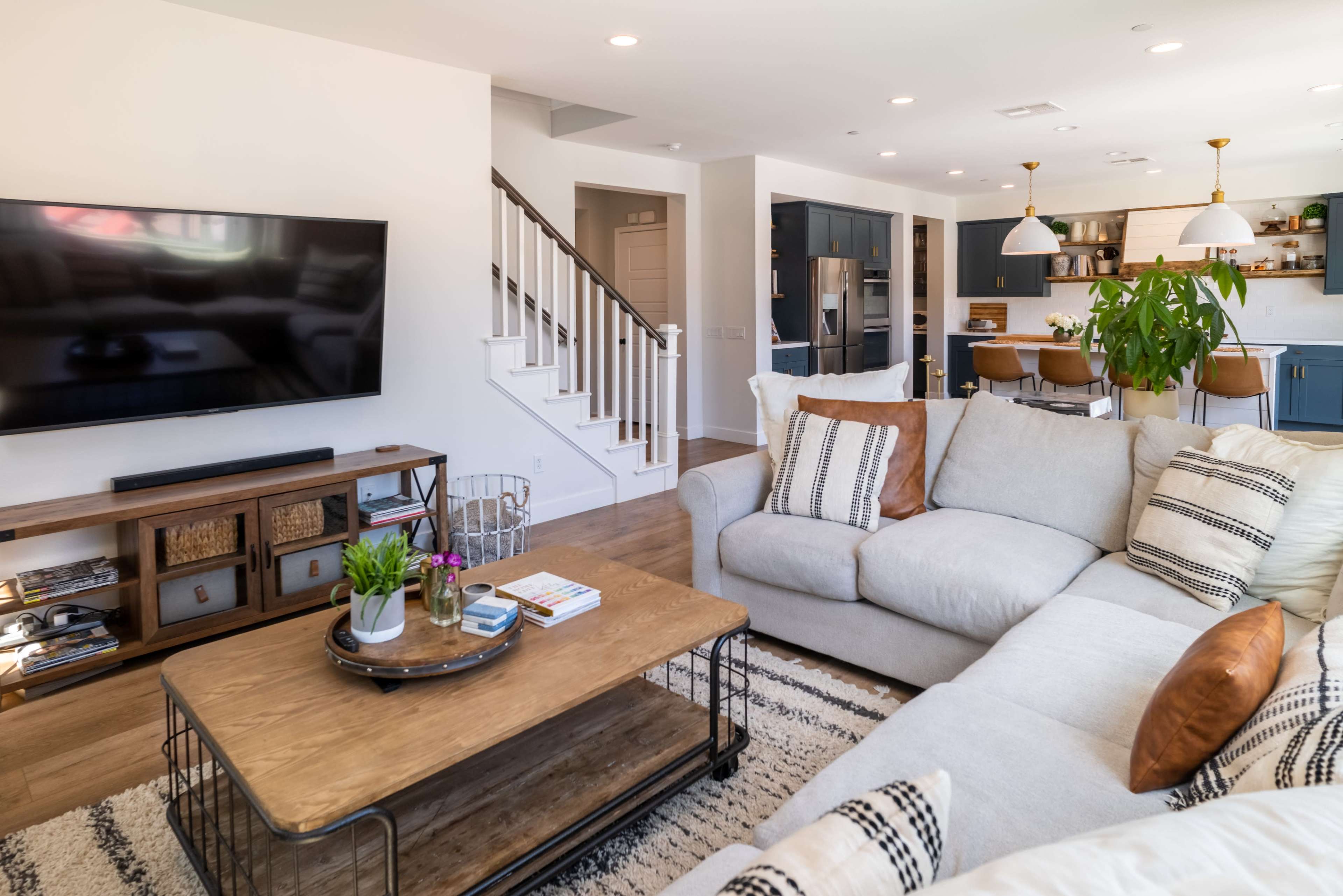 A modern living room features a large sectional sofa, a coffee table, a flat-screen TV mounted on the wall, and a dining area in the background.