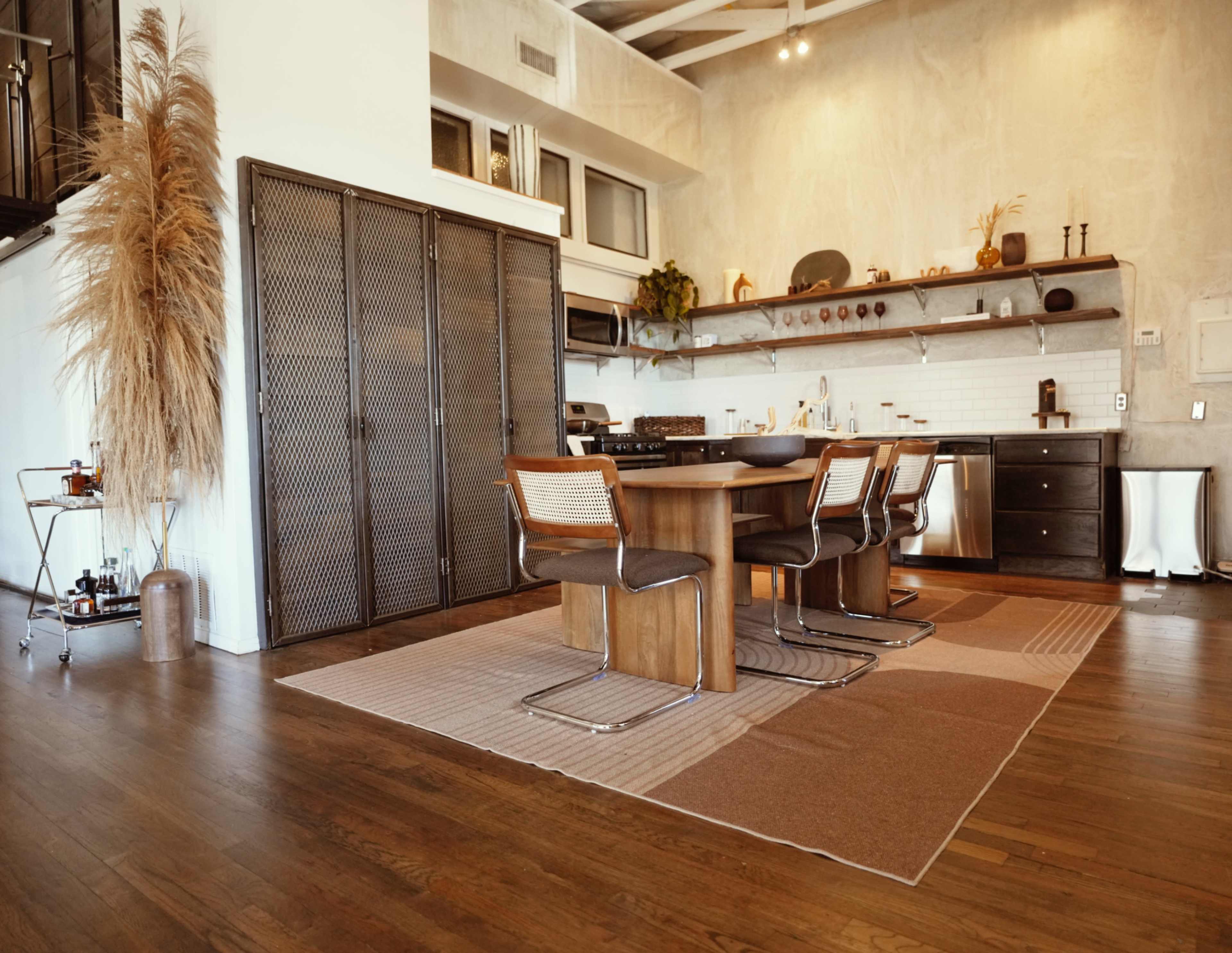The image shows a modern kitchen with a wooden dining table and metal chairs, accented by large decorative plants and open shelving.