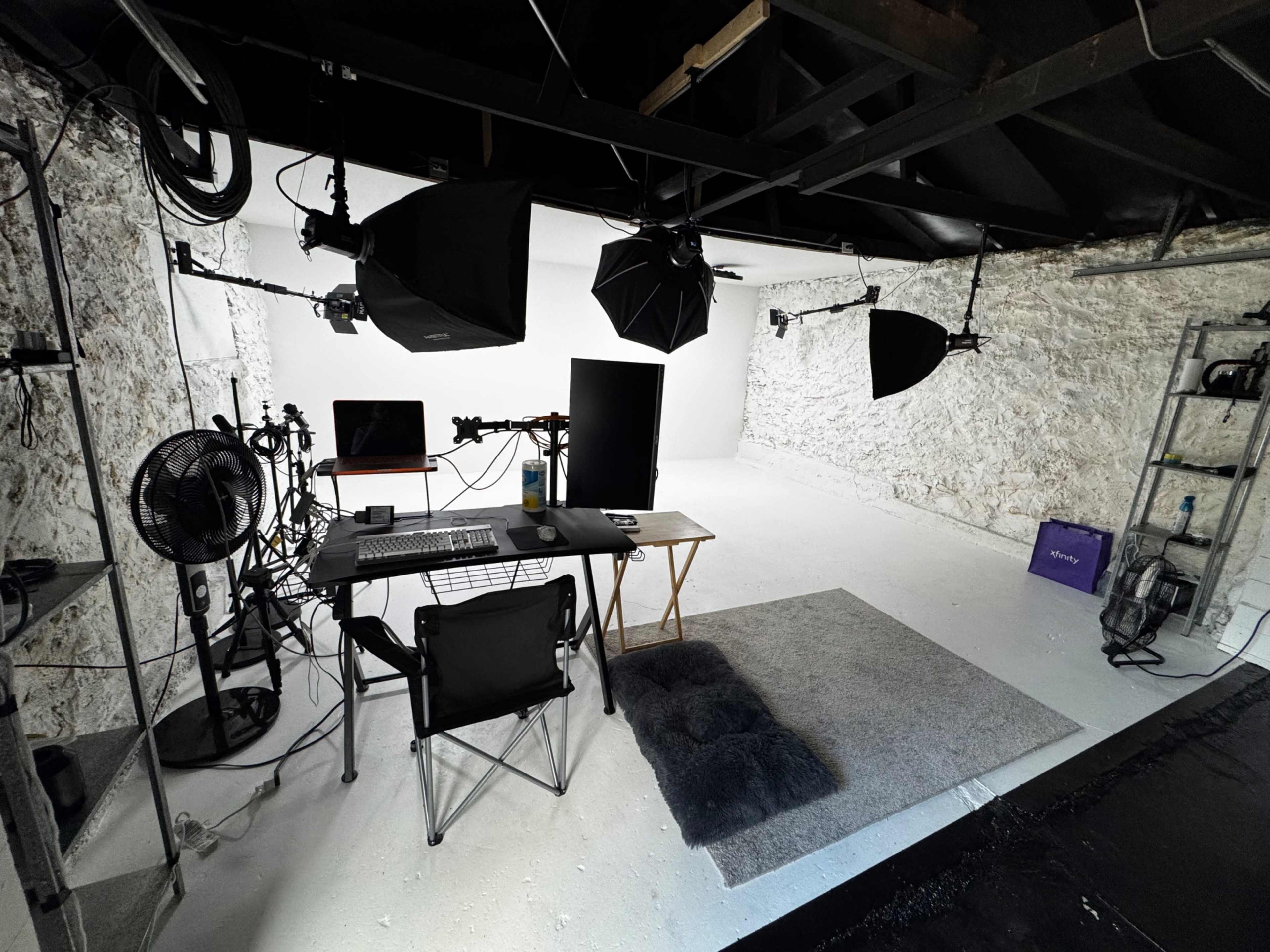 The image shows a minimalistic photo studio setup with a desk, computer, and studio lights against a plain white backdrop.