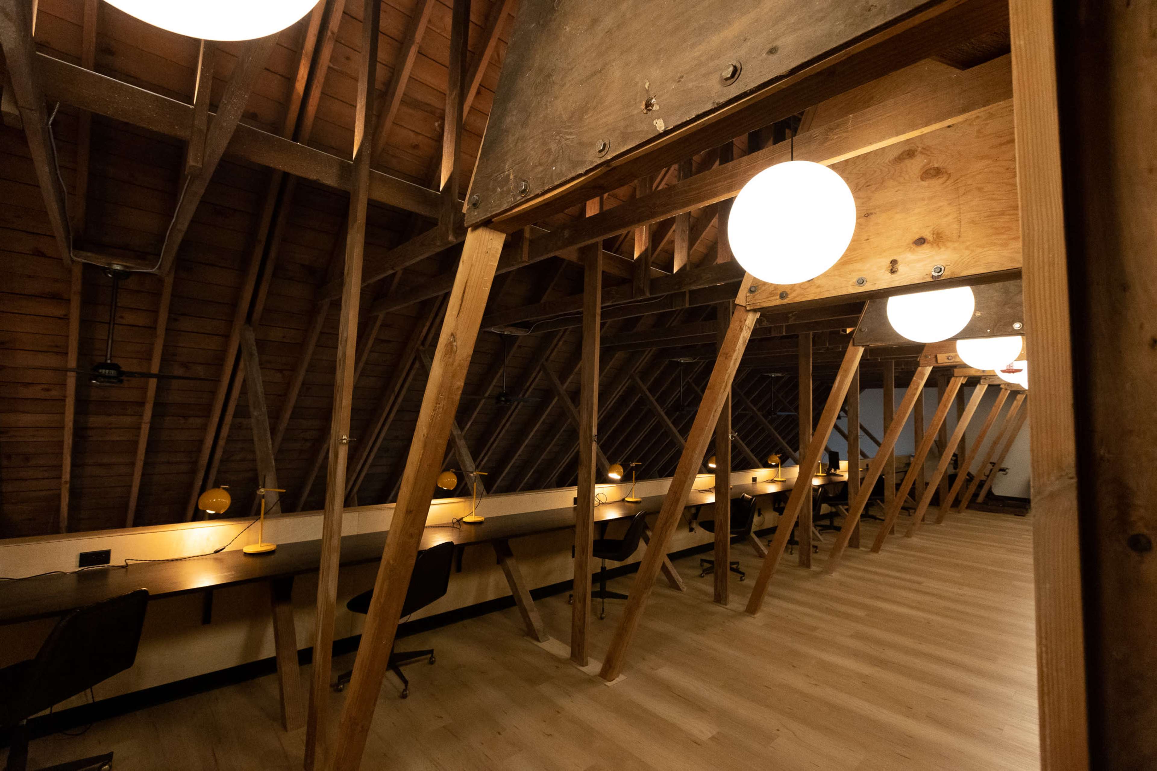 The image shows an attic space with wooden beams and pendant lights, featuring a row of desks and chairs against the walls.