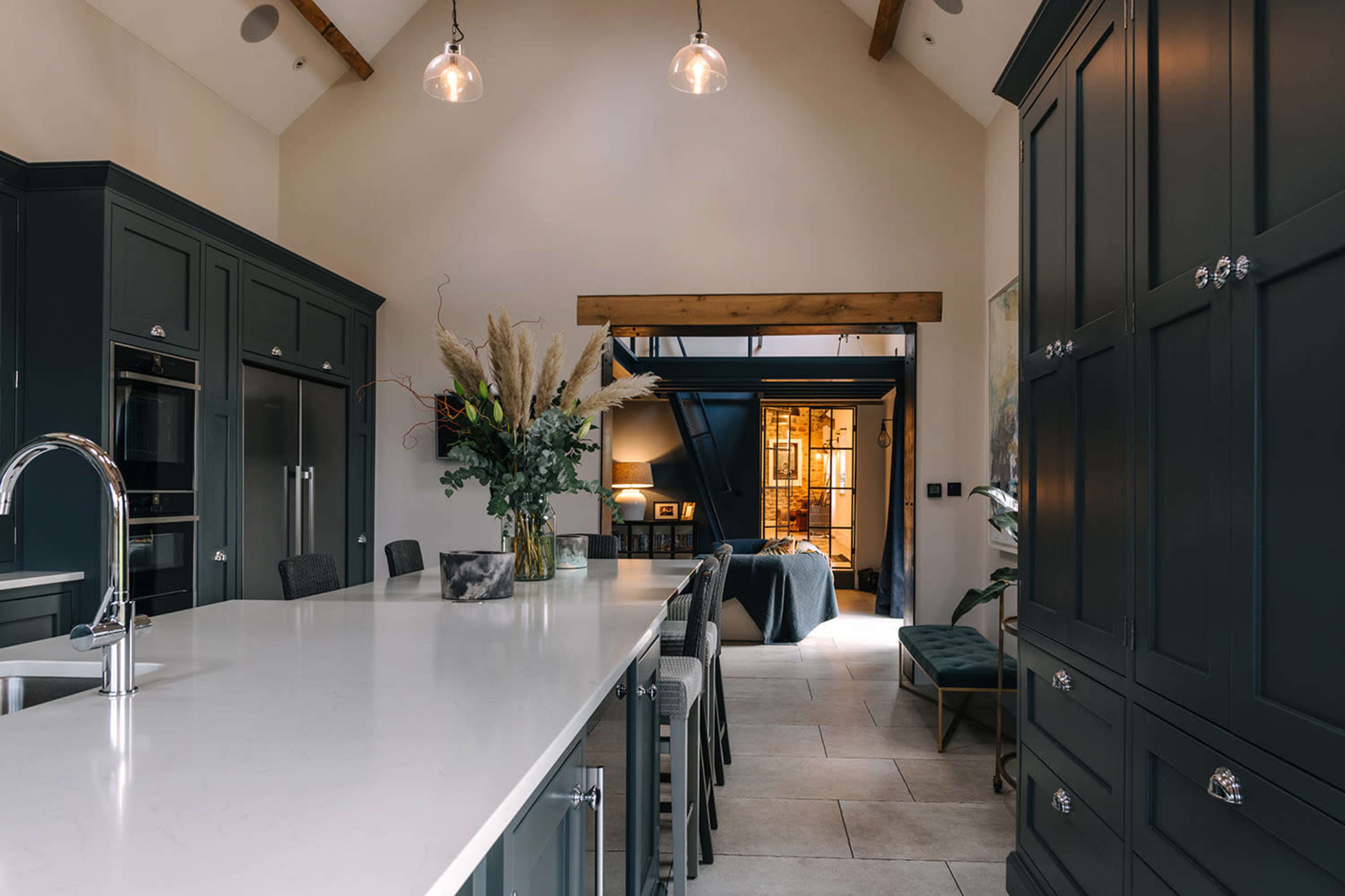 A modern kitchen features a long countertop with stools, dark cabinetry, and a view into an adjacent living space with natural light.